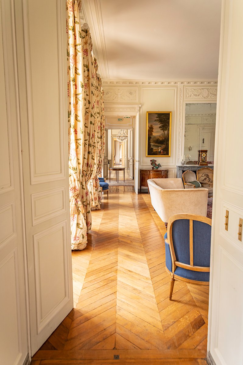 A view down the enfilade shows the  living room with a polished wooden floor, large windows, pretty curtains and a painting.