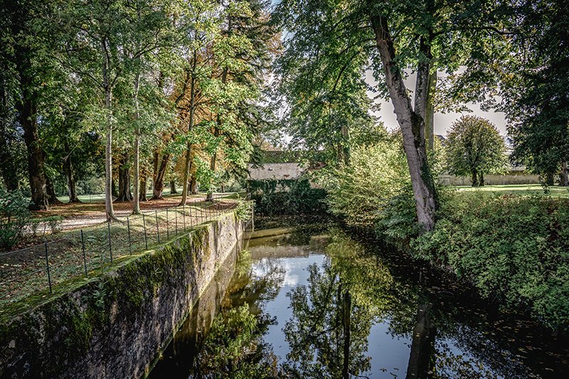 A moat winds through a grassy area with trees lining its banks on the grounds of Chateau de Courtomer.