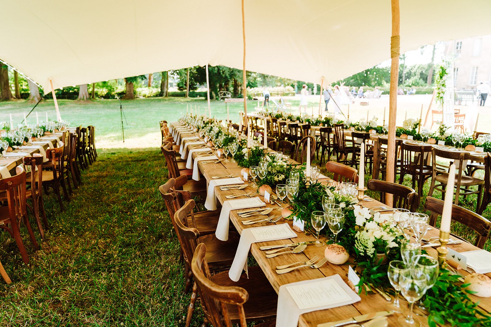 Long tables setup under a marquee with wooden chairs and decorated with flowers for a wedding at the chateau.