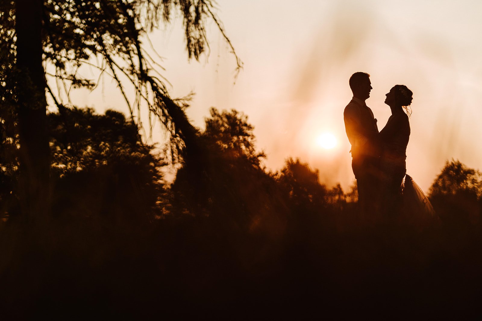 A silhouette of a bride and groom standing in a field at sunset, with a french chateau wedding venue in the background.