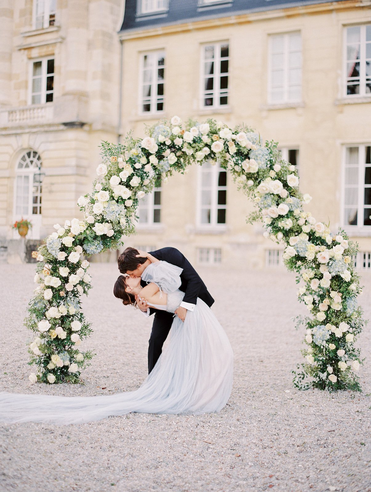 A bride and groom kissing under a floral arch in front of Chateau de Courtomer, a French chateau wedding venue near to Paris.