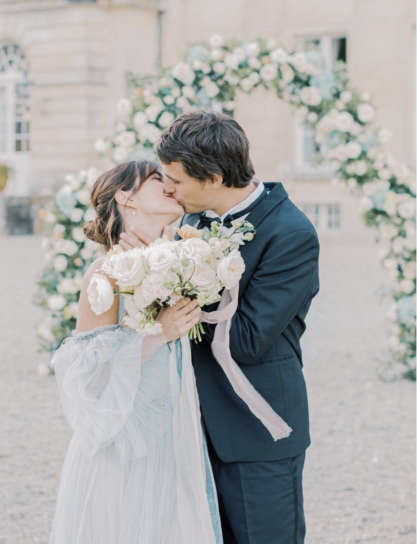 A bride and groom kissing under a flower wedding arch in front of Chateau de Courtomer, an ideal wedding venue.