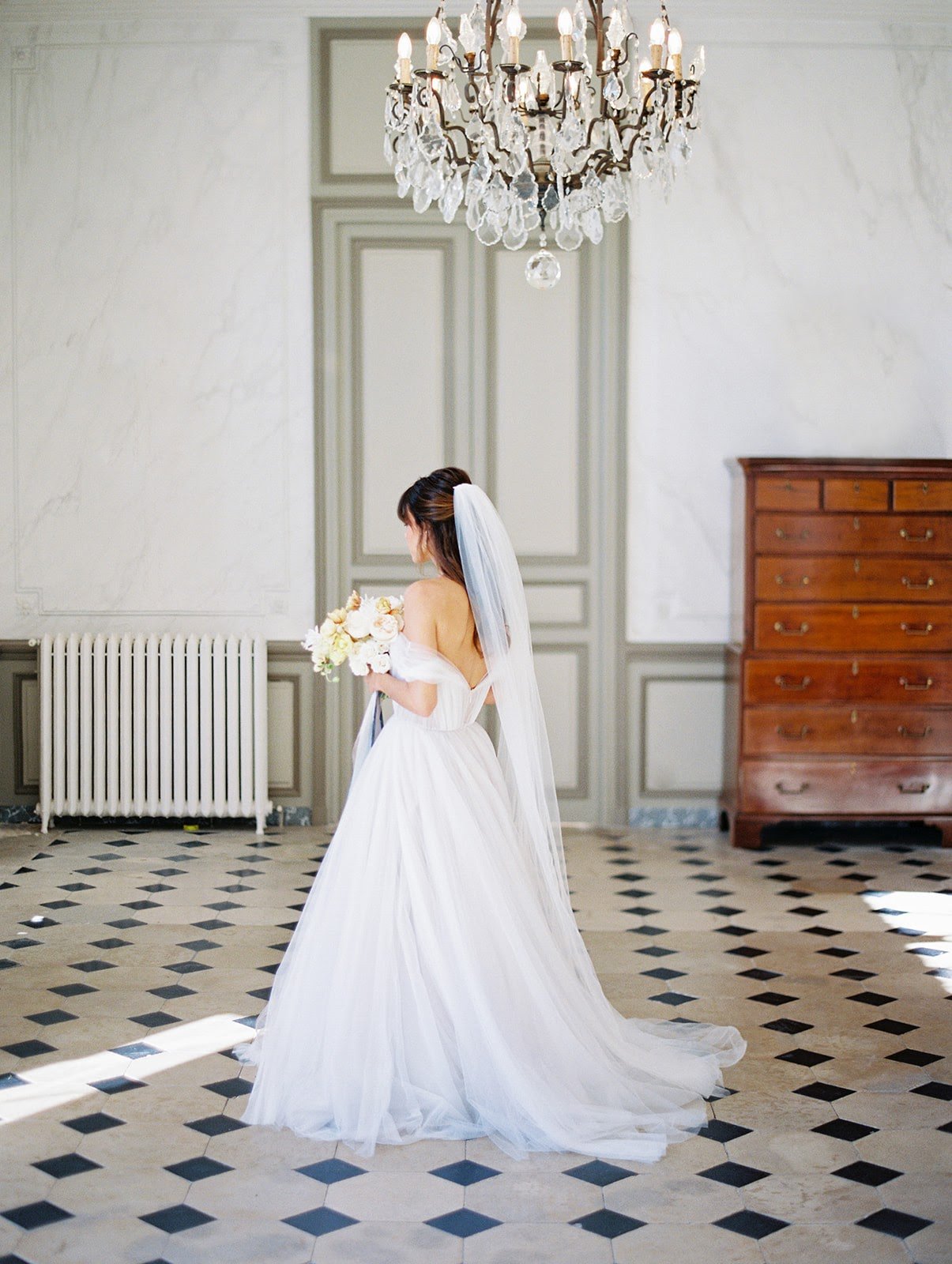 A bride, standing under a glittering chandelier in the entrance hall of Cde Courtomer, the perfect venue for a wedding.