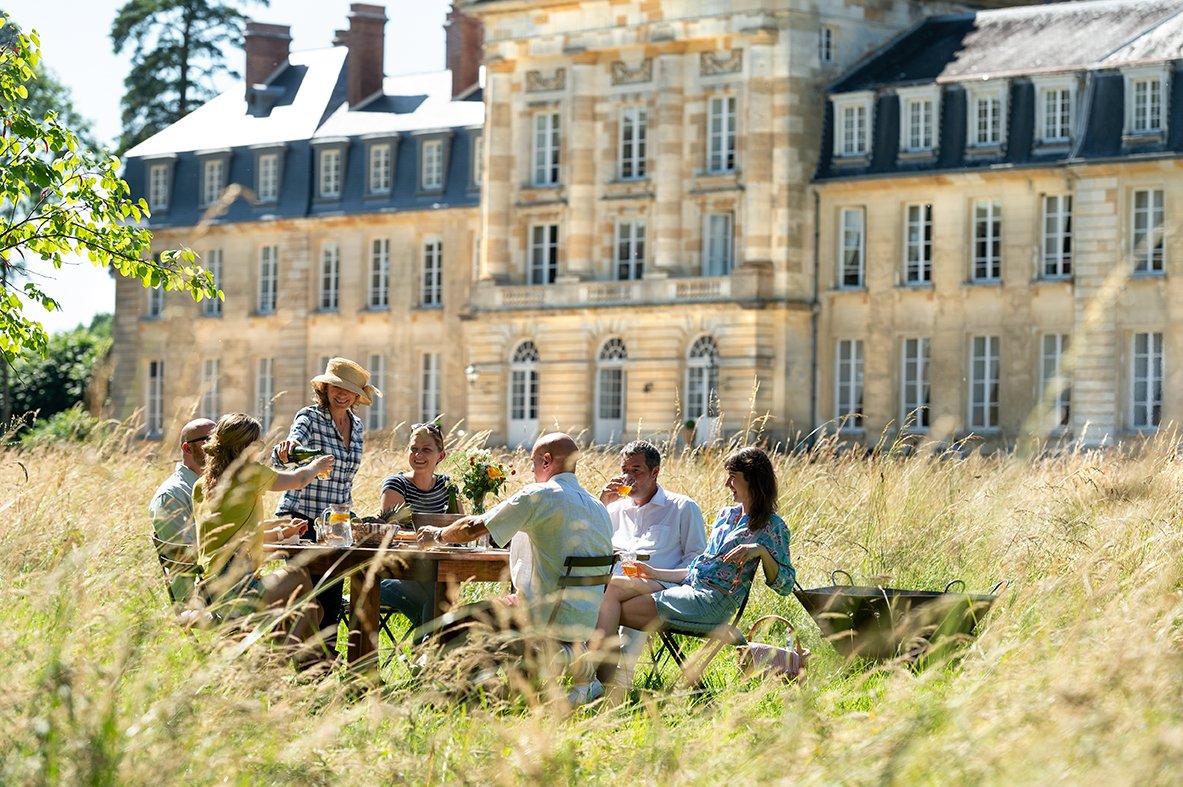 A group of people picnicing at a table in the grass relaxing in front of Chateau de Courtomer, a unique place to stay on holiday.