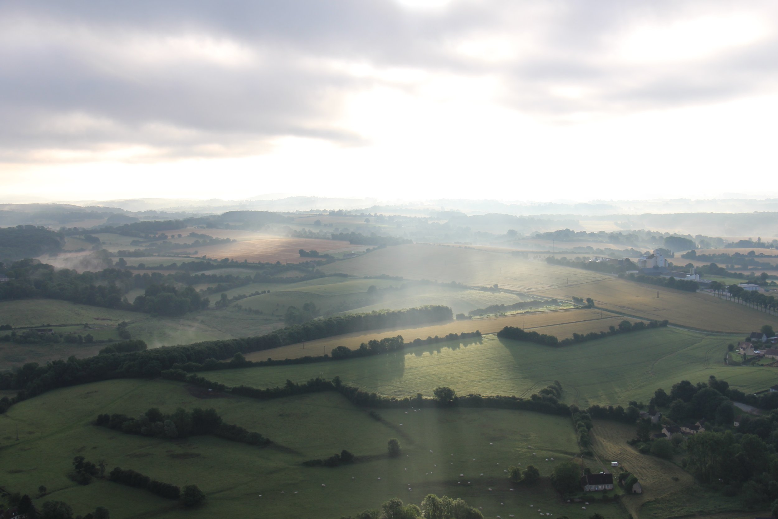 An aerial view of lush green fields and farmland, with old french farm buildings and a cloudy sky.