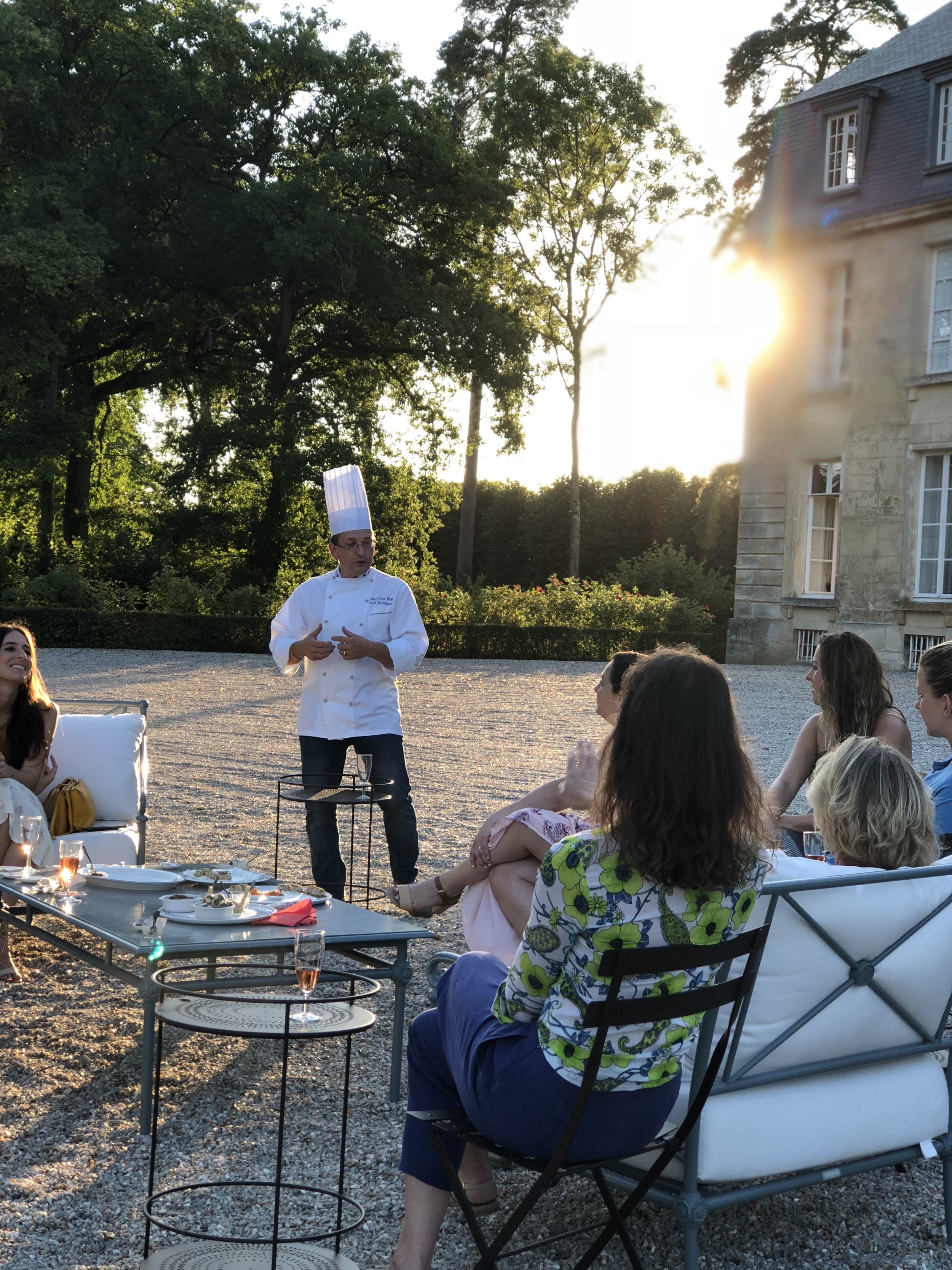 People relaxing with a drink and food on comfortable chairs outside the chateau at sunset, listening to a chef talking.