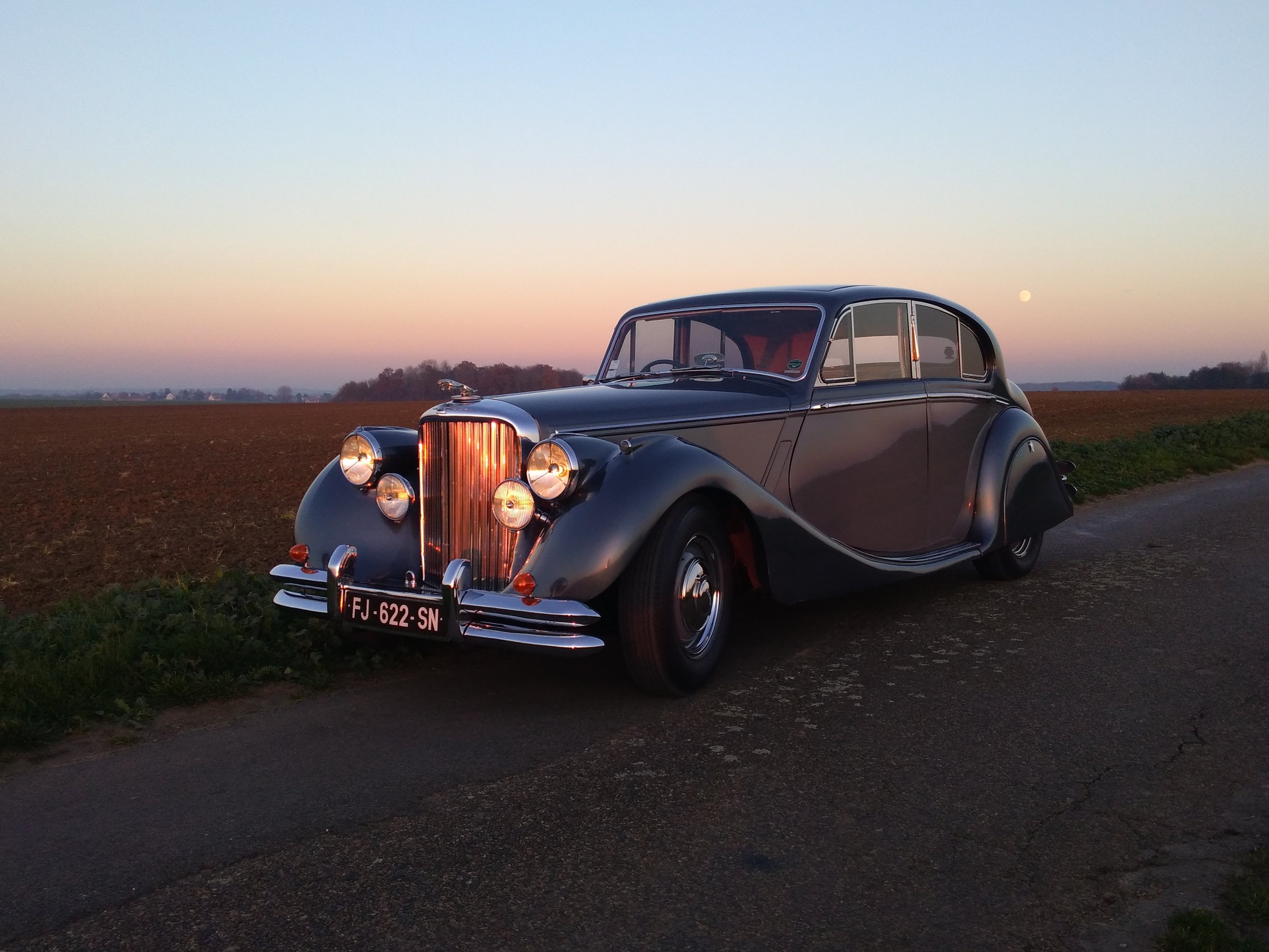A classic car is parked on a country road at sunset near Chateau de Courtomer.