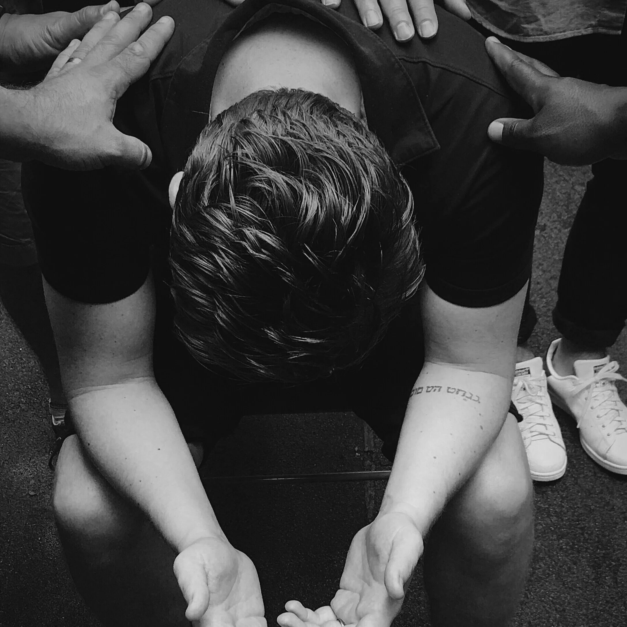 A person sitting with their head bowed, arms resting on their knees, with a tattoo on their right arm. Three hands are placed on their shoulders and head, suggesting support or comfort. The photo is in black and white.