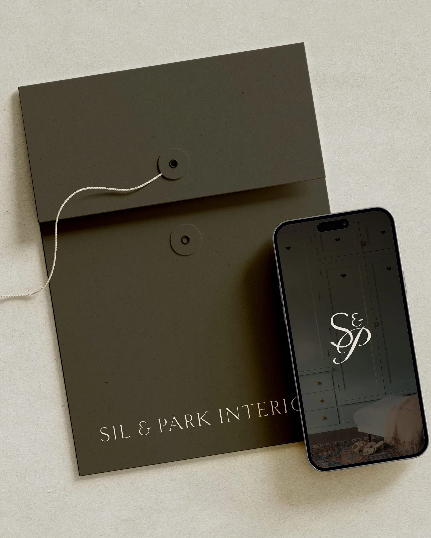A dark green folder for SIL & PARK Interiors with a string. Next to the folder is a smartphone displaying the SIL & PARK Interiors logo icon. Created by Clarke Creative.