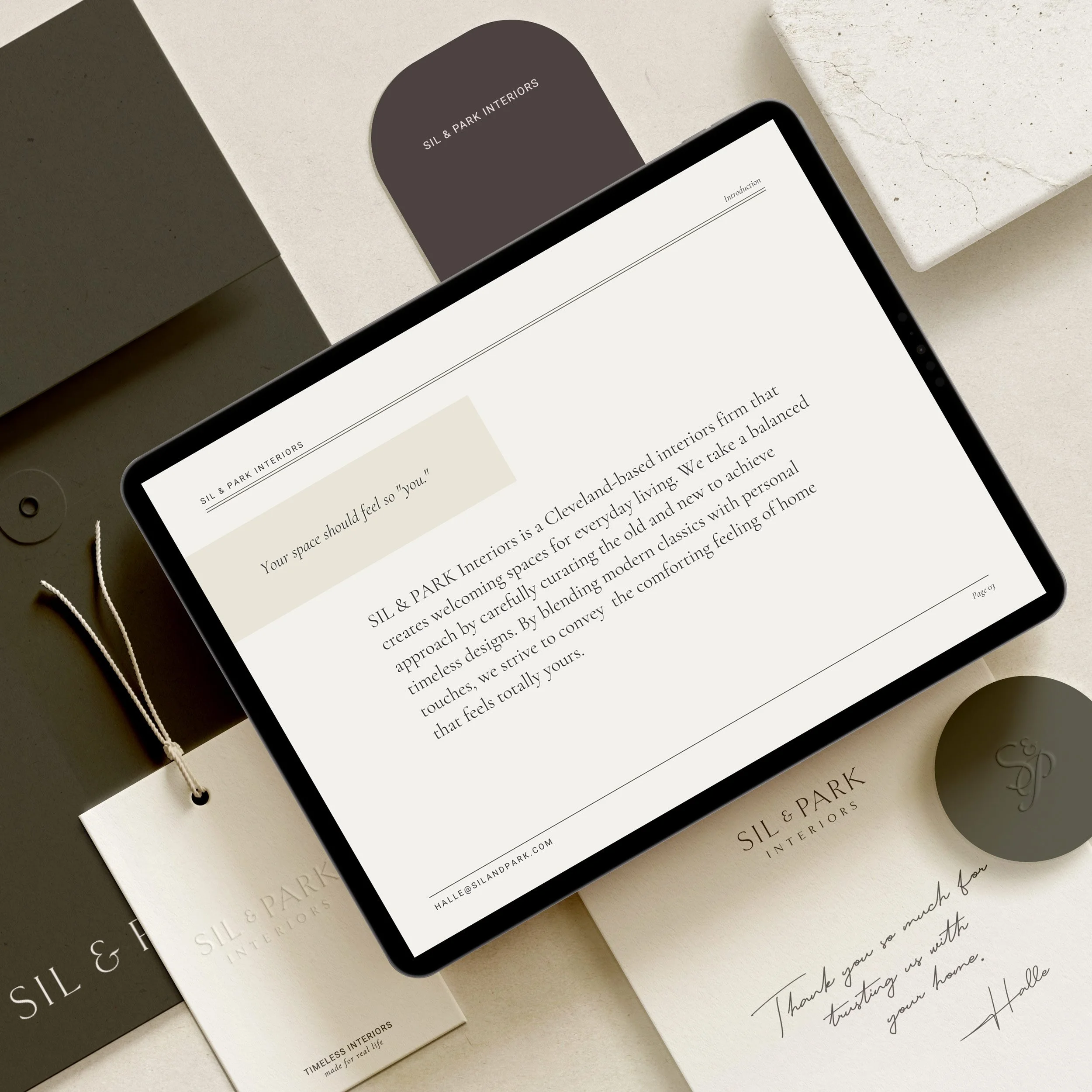 Digital tablet displaying a welcome message from SIL & PARK Interiors, surrounded by branding materials including a tag, a card, and a booklet, all in neutral tones. Created by Clarke Creative.