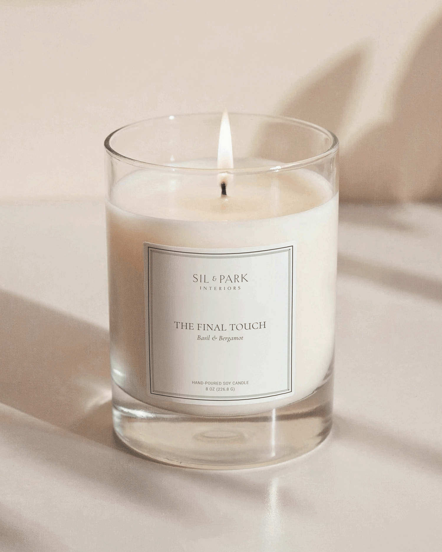 A lit candle in a glass holder labeled 'Sil & Park Interiors, The Final Touch, Basil & Bergamot, Hand-Poured Soy Candle, 8 oz (226.8 g)' on a light-colored surface. Created by Clarke Creative.