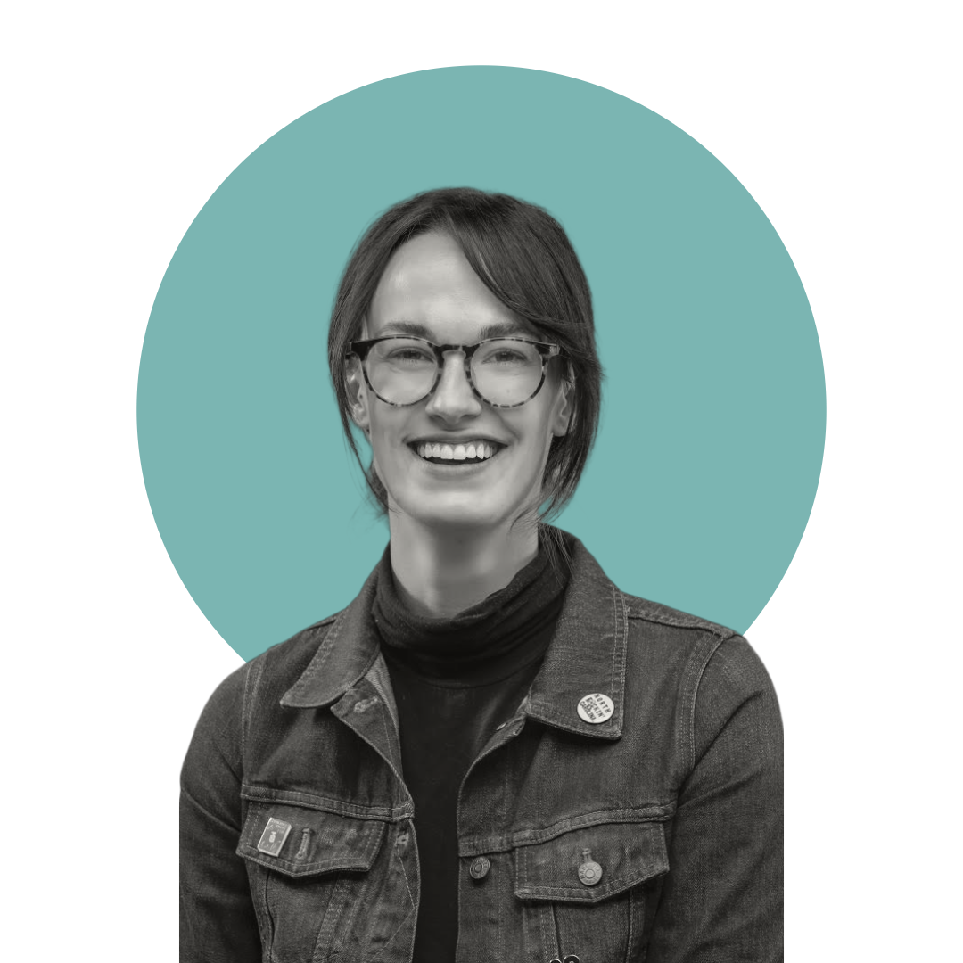 A black and white portrait of a smiling woman with short hair, glasses, a denim jacket, and a pin on her lapel, set against a teal circular background.