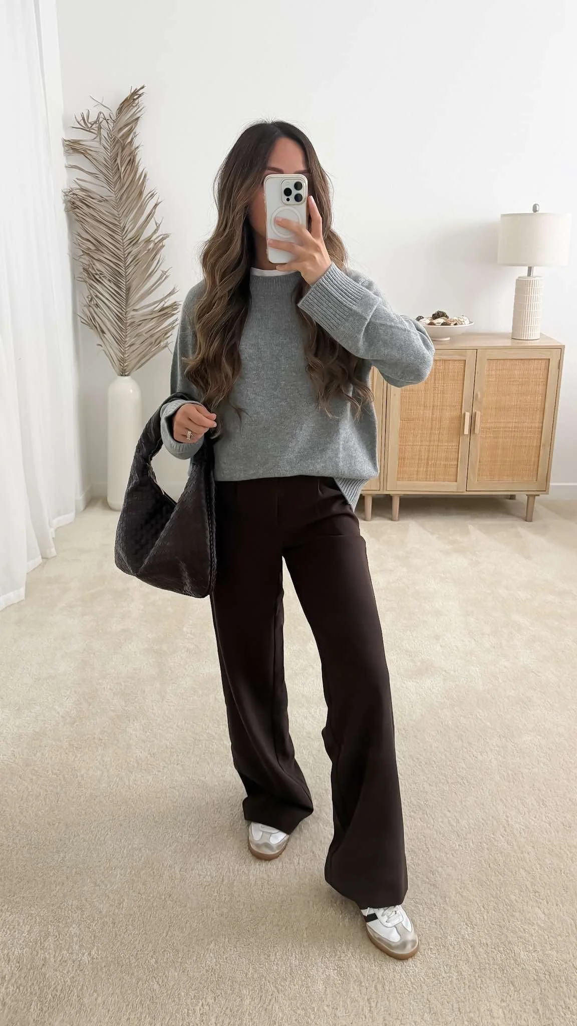 Dark brown trousers outfit with a gray sweater layered over a white t-shirt, neutral sneakers, and dark brown woven bag