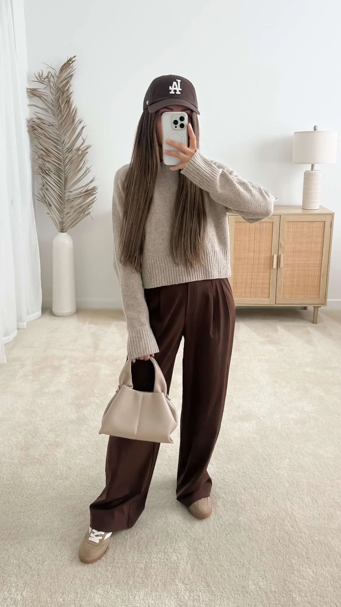 Brown pants outfit styled with brown tailored pants, a neutral sweater, brown sneakers, beige handbag and brown baseball cap