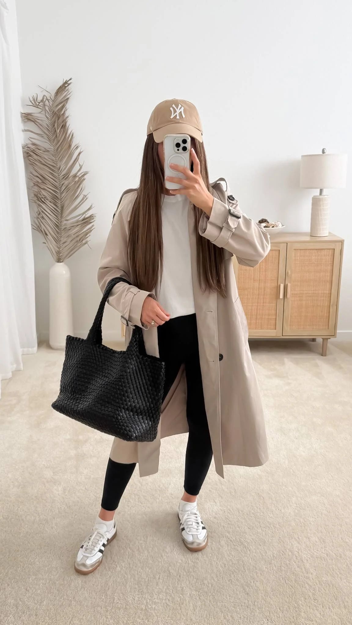 White T-shirt styled with a beige trench coat, black leggings, white sneakers, and a black woven tote bag.