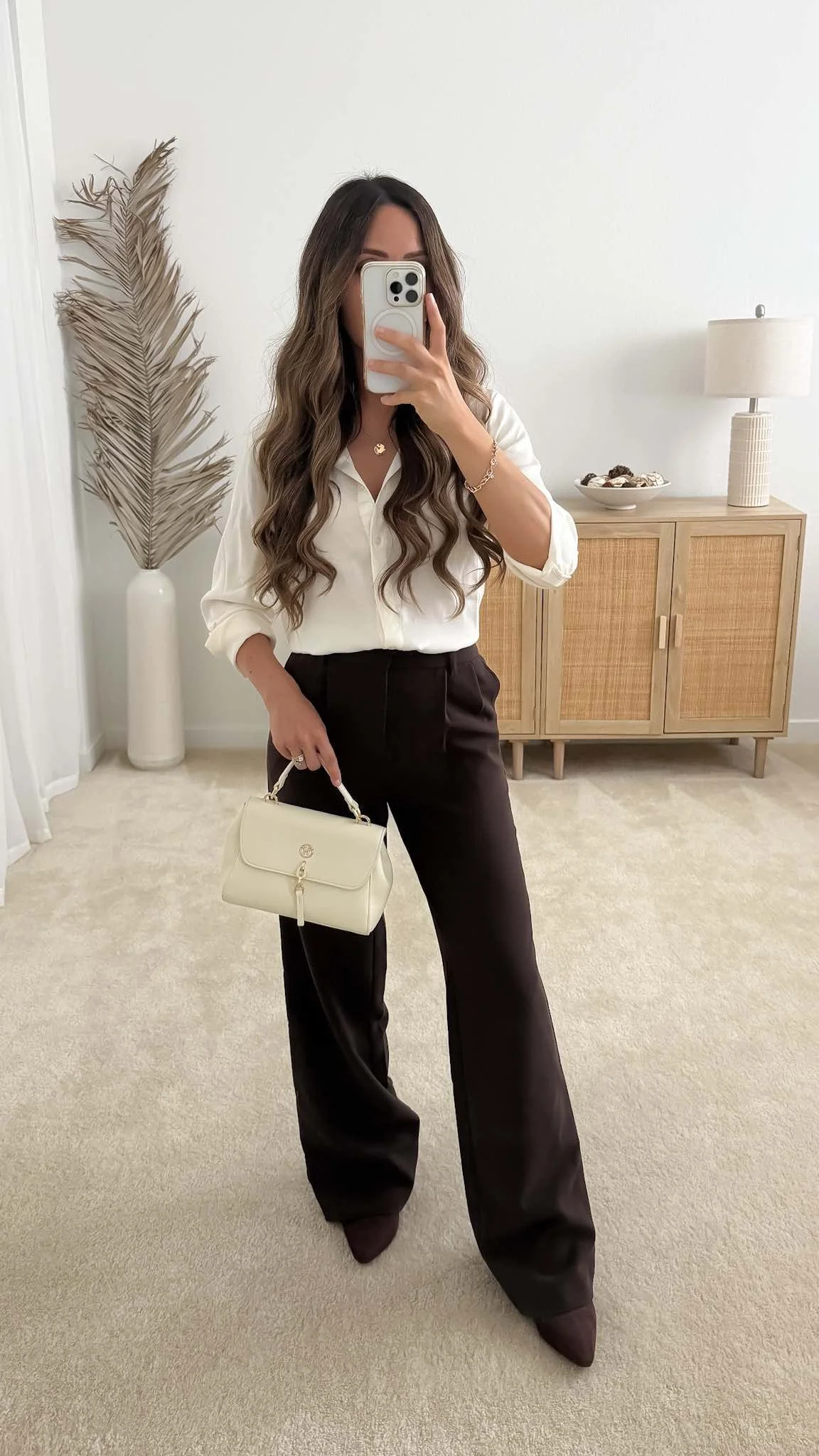 Dark brown trousers outfit with an ivory blouse, brown ankle boots, and cream handbag