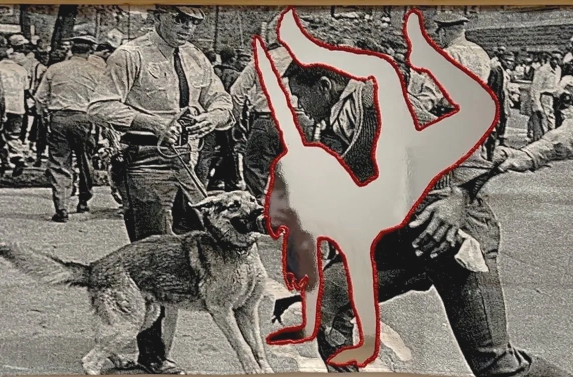 A black & white photo printed onto cloth. An image of a unicorn is cut out from the cloth and backed with mirrored acrylic. The unicorn is battling with a dog during the Birmingham Confrontation 1963.
