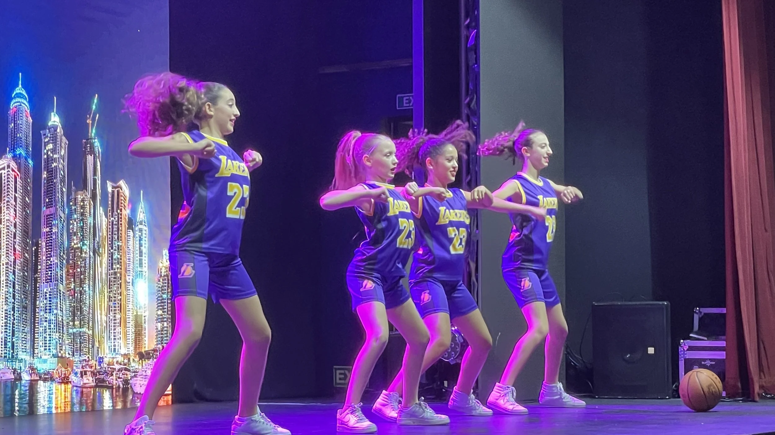 Five young girls in blue and yellow basketball uniforms performing a dance routine on stage, with a cityscape backdrop and a basketball on the floor to the right.