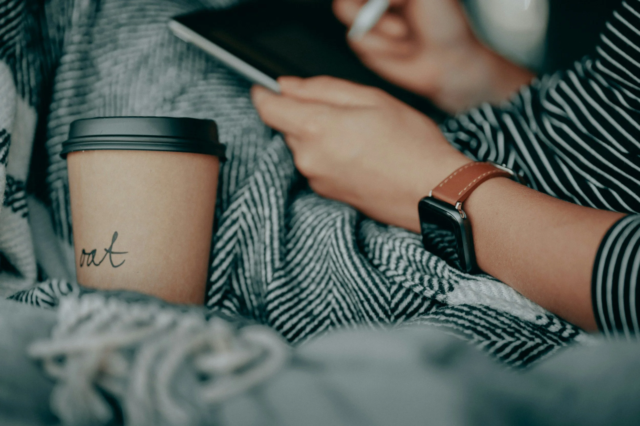 Person holding a smartphone, wearing a striped shirt and a smartwatch, with a paper coffee cup and a mug on a cozy blanket.
