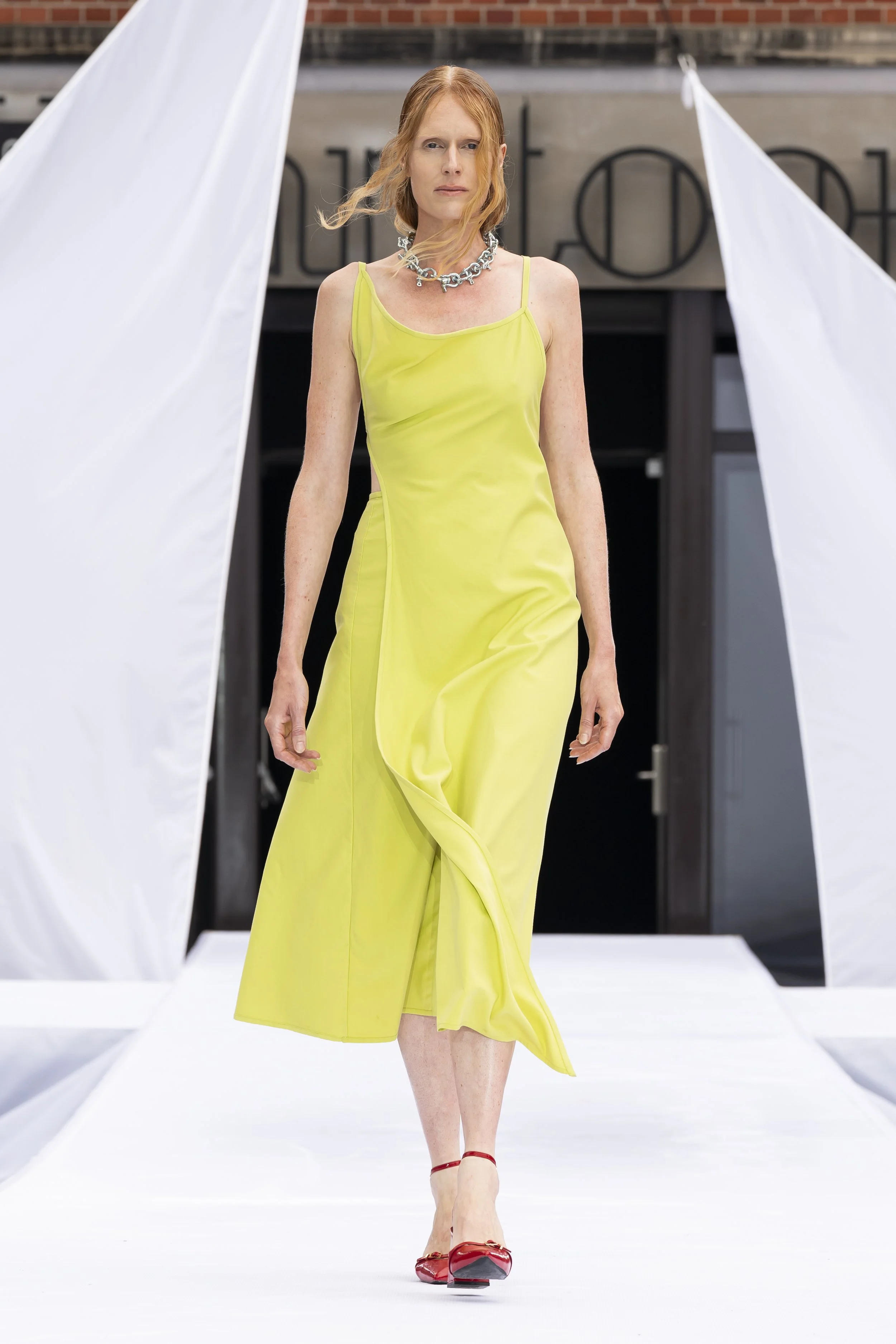 A woman with red hair walking on a runway during a fashion show, wearing red high heels, a chunky silver necklace and a yellow satin slip dress by Martan, a circular fashion brand. Actress, writer & creative developer Jolanda van den Berg.