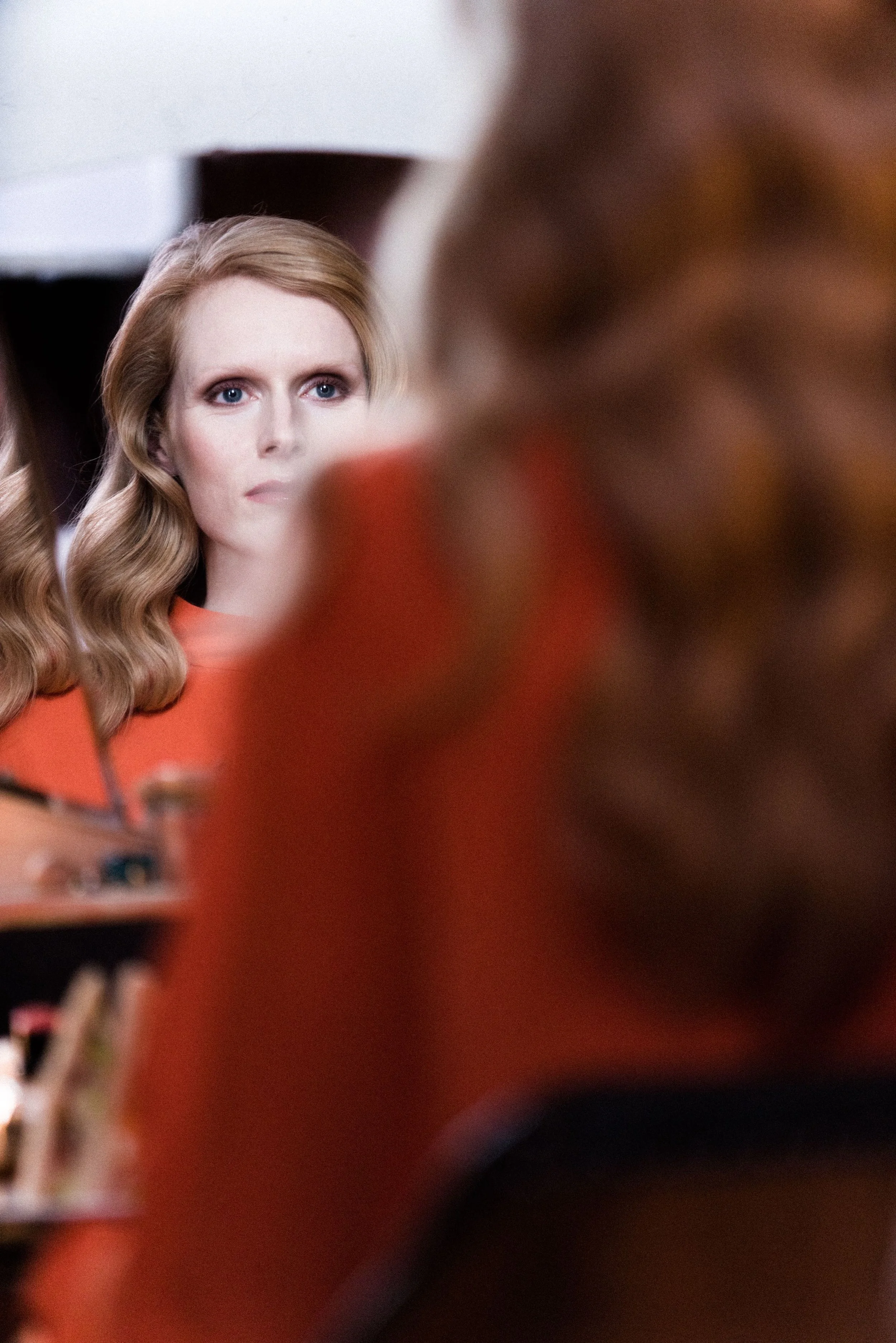 A woman with red hair and blue eyes looking into a mirror of a vintage cabinet. Actress, writer & creative developer Jolanda van den Berg.