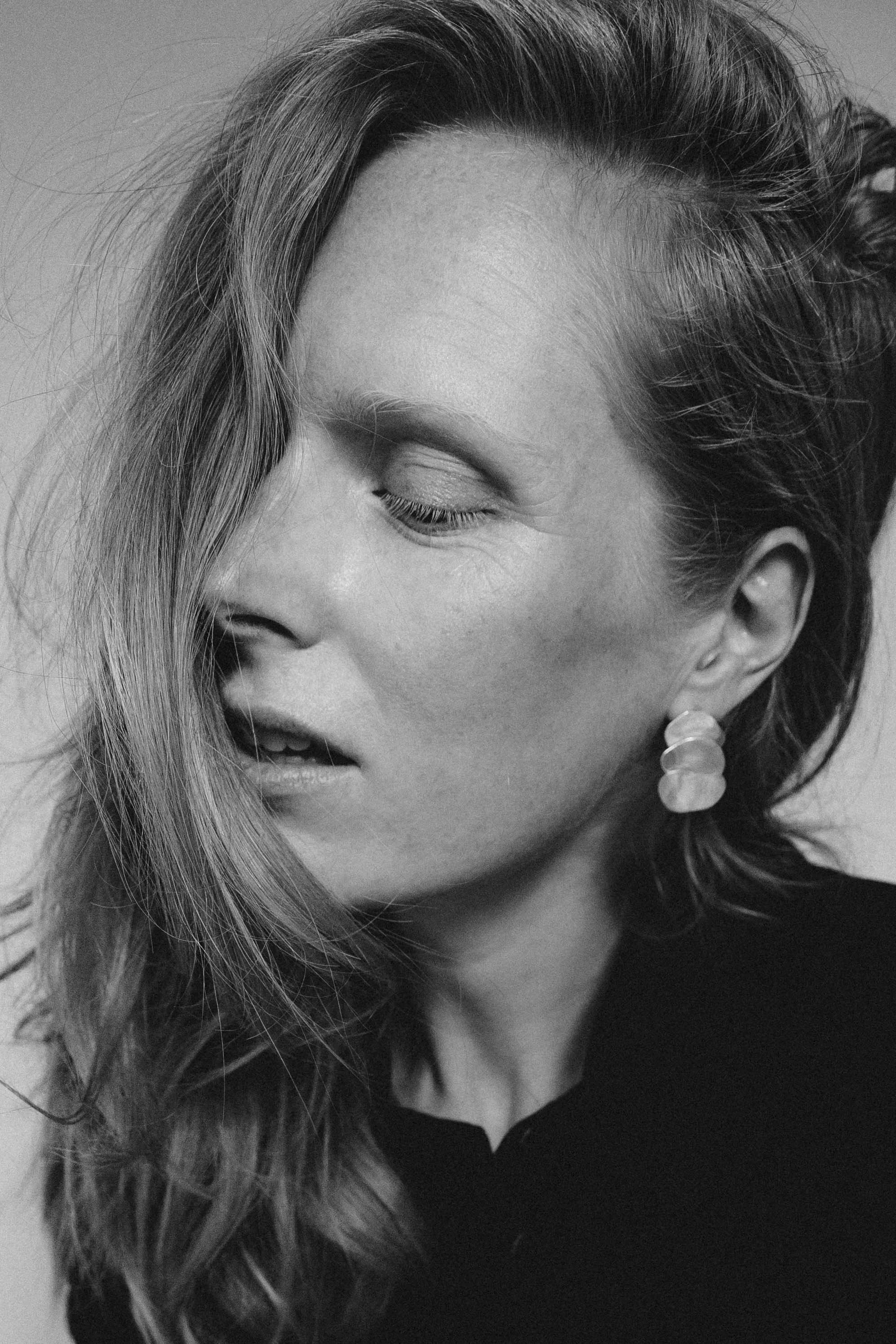 A close-up black and white photo of a woman with wavy hair and earrings, with her eyes closed and her mouth slightly open, looking down. Actress, writer & creative developer Jolanda van den Berg. Photography by Chloé Chanté Leenheer.