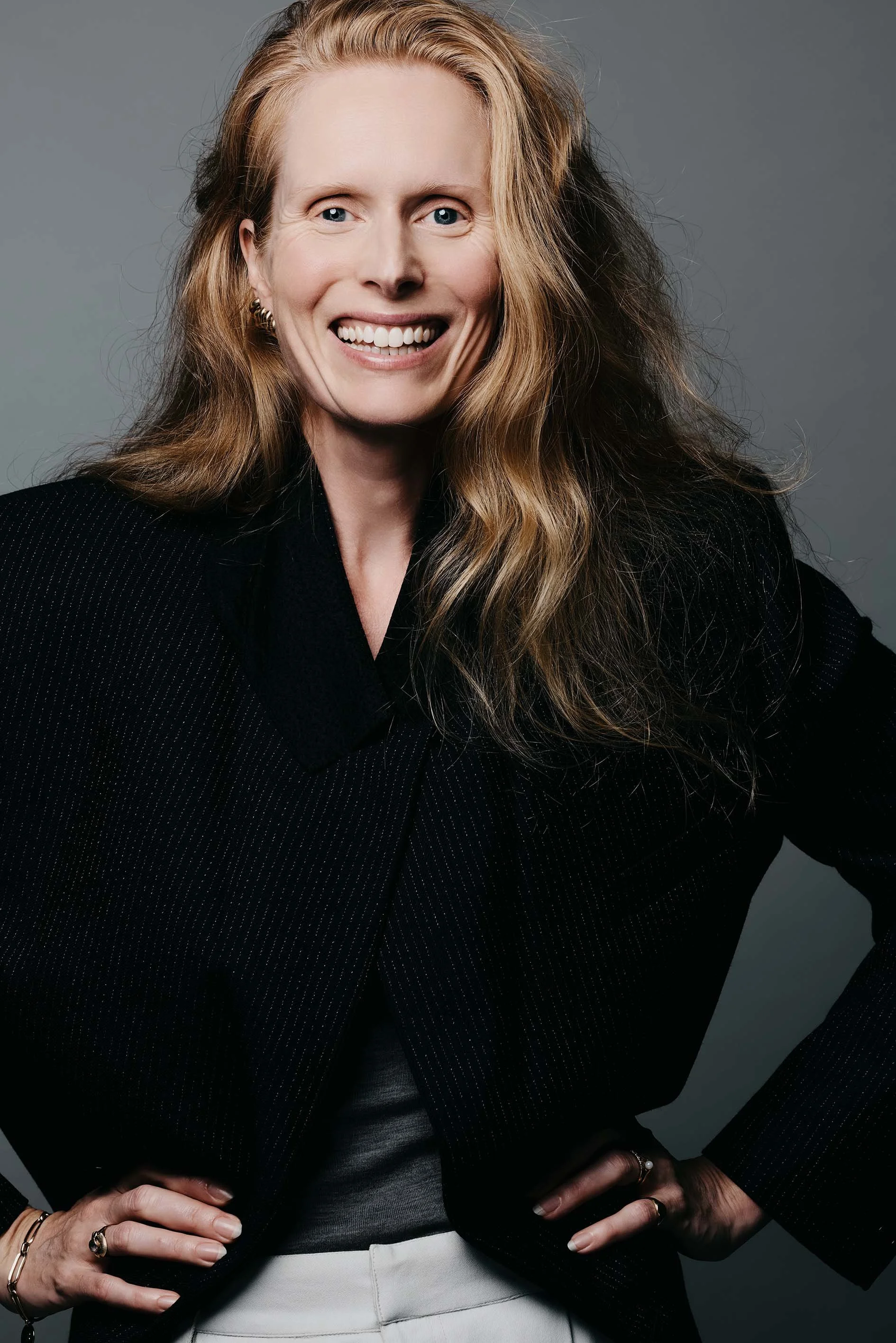 Professional headshot of Jolanda van den Berg, actress, smiling warmly with long auburn hair, wearing a black pinstriped blazer by Dutch brand Róhe against a gray background. Photo credit: Anne van Zantwijk.