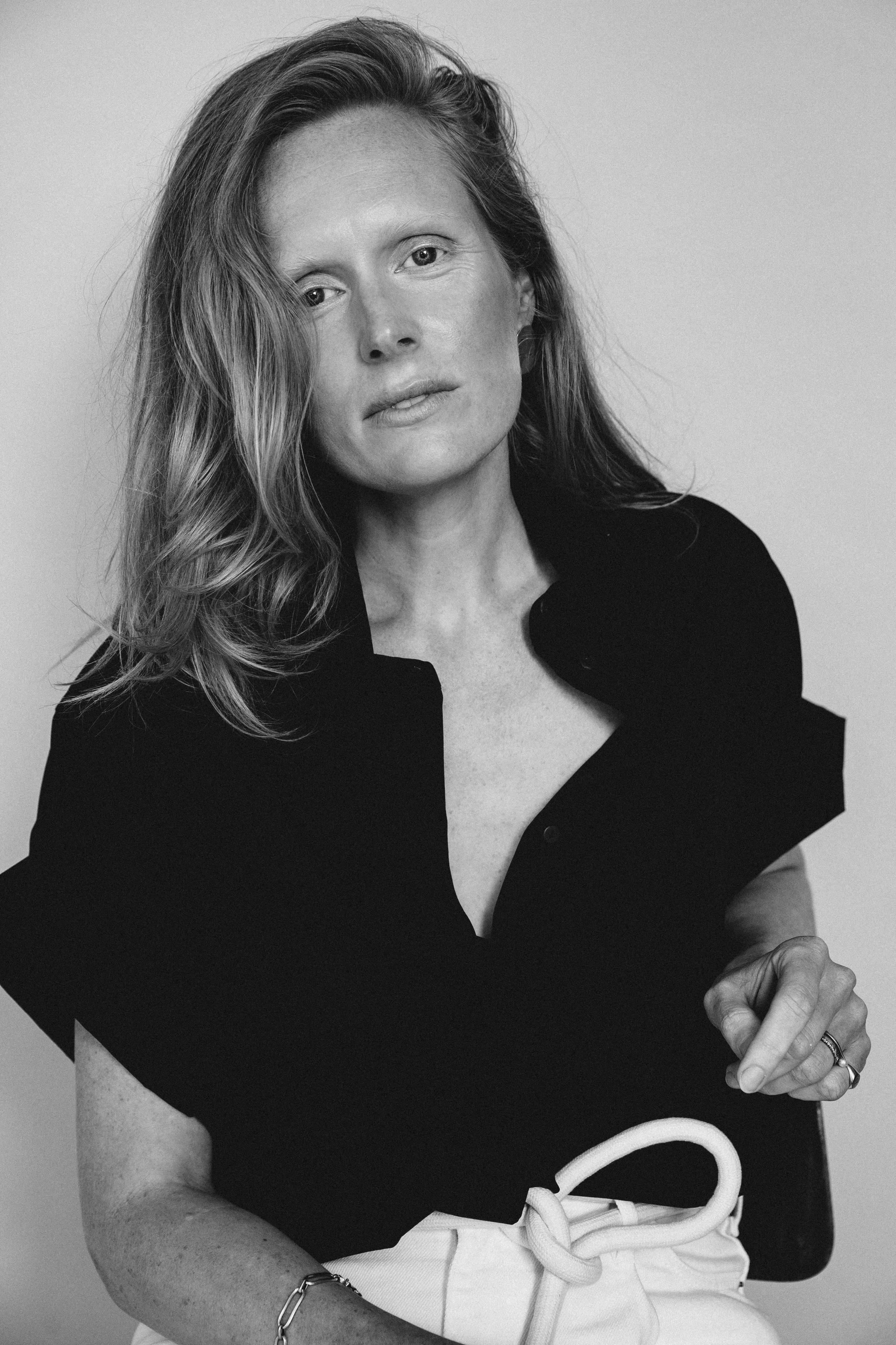 Black and white portrait of a woman with wavy hair, wearing a dark top and light-colored pants with a belt, looking directly at the camera. Actress, writer & creative developer Jolanda van den Berg. Photography by Chloé Chanté Leenheer.