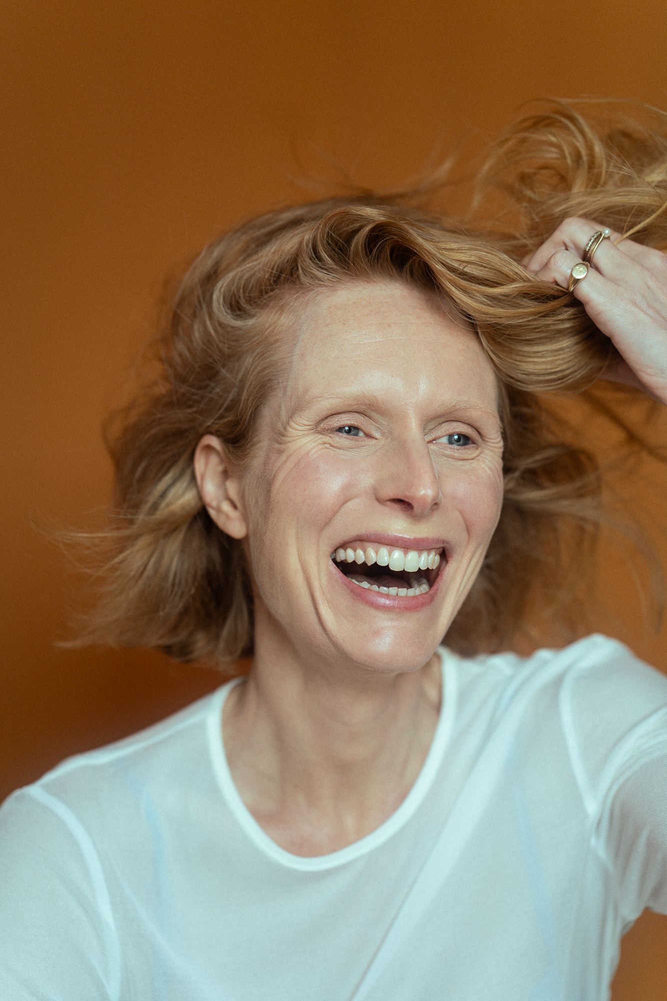A woman with red hair laughing, holding her hair with one hand, wearing a white shirt, and standing in front of a brown background. Actress, writer & creative developer Jolanda van den Berg. Photography by Anna Perger.