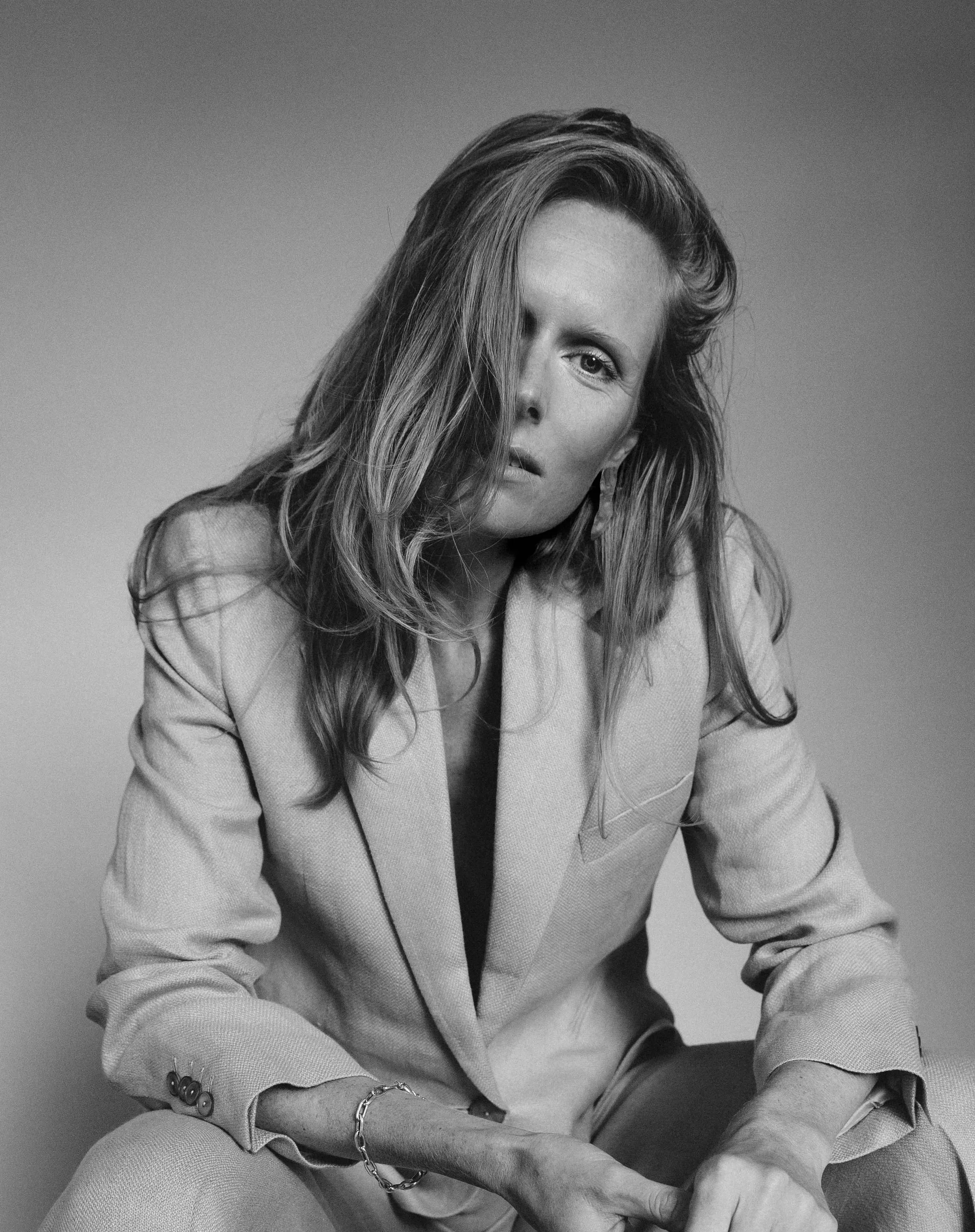 Black and white portrait of a woman with tousled hair, wearing a light-colored blazer and pants, sitting with one leg crossed, looking at the camera with a serious expression. Actress, writer & creative developer Jolanda van den Berg. Photography by 