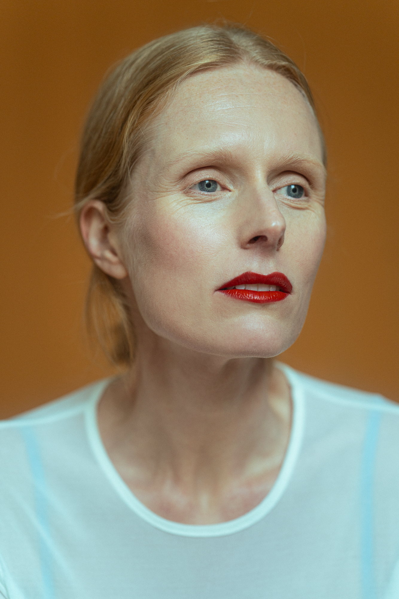 Close-up of a woman with minimal makeup and red lipstick and light blue eyes, wearing a white shirt with blue stripes, against an orange background. Actress, writer & creative developer Jolanda van den Berg. Photography by Anna Perger.
