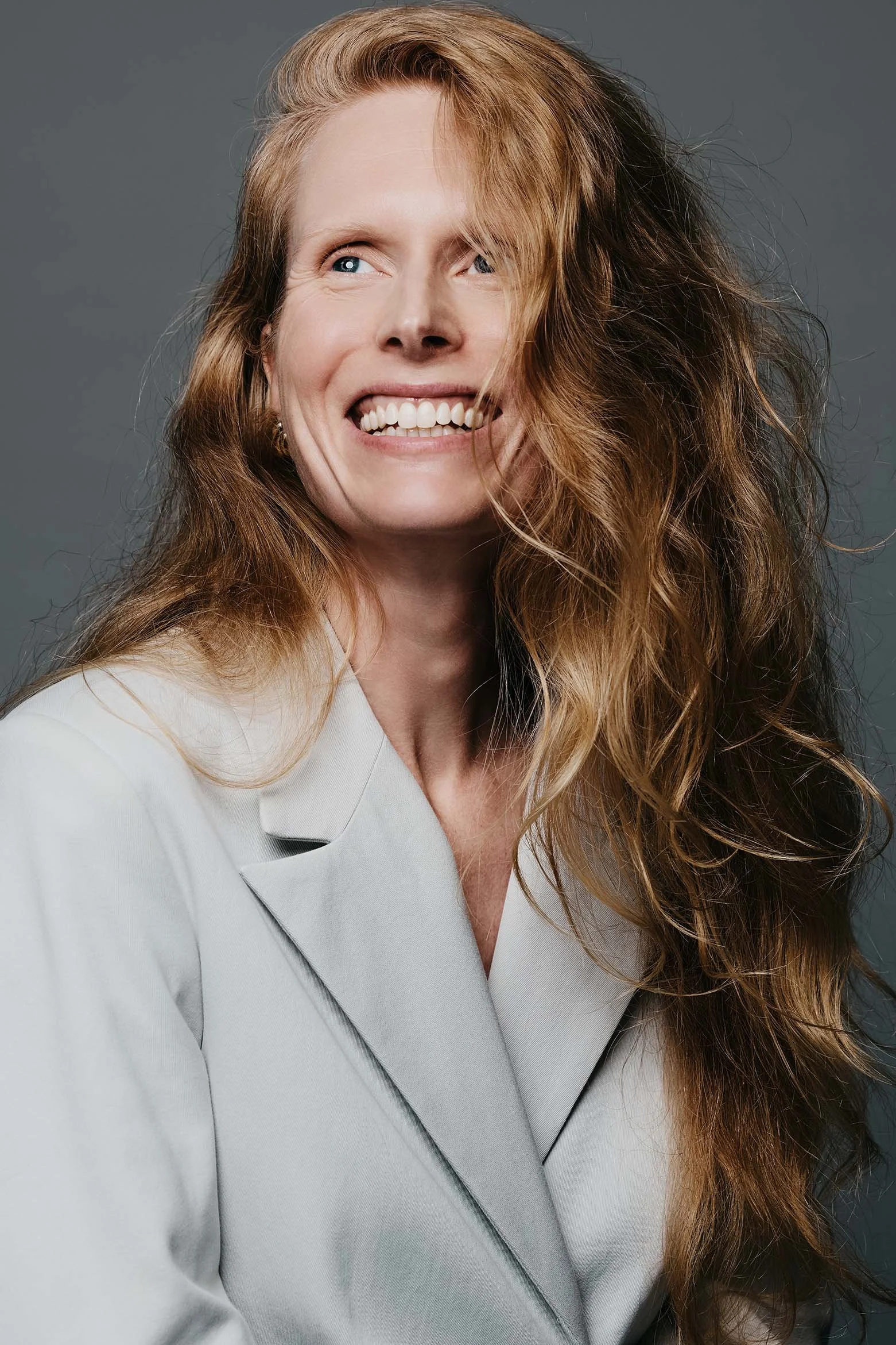 Professional headshot of Jolanda van den Berg, actress, smiling warmly with long auburn hair, wearing a light gray blazer against a gray background. Photo credit: Anne van Zantwijk.