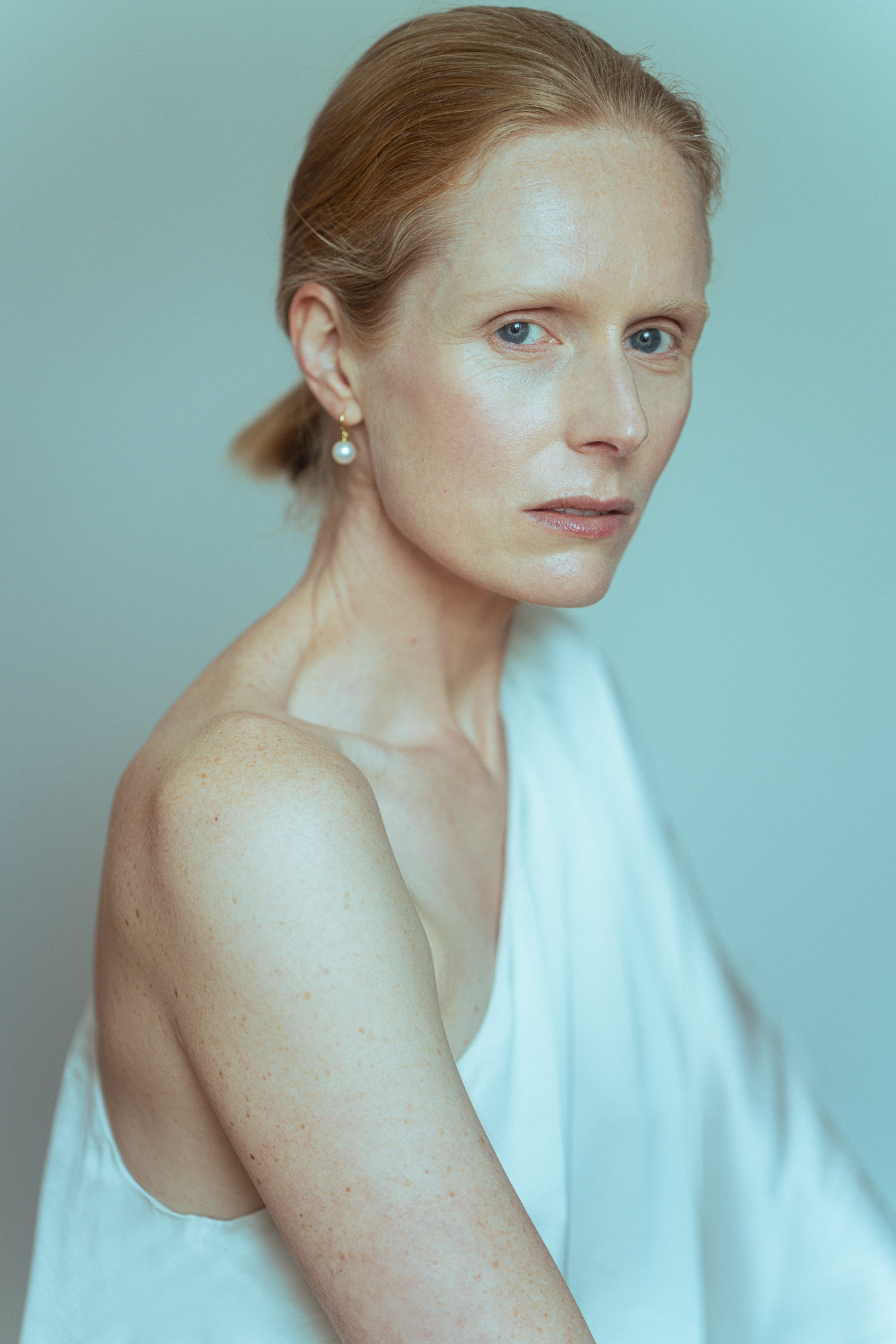 A woman with red hair wearing a pearl earring a white sleeveless top and minimal makeup, looking at the camera with a neutral expression against a plain background. Actress, writer & creative developer Jolanda van den Berg. Photography by Anna Perger
