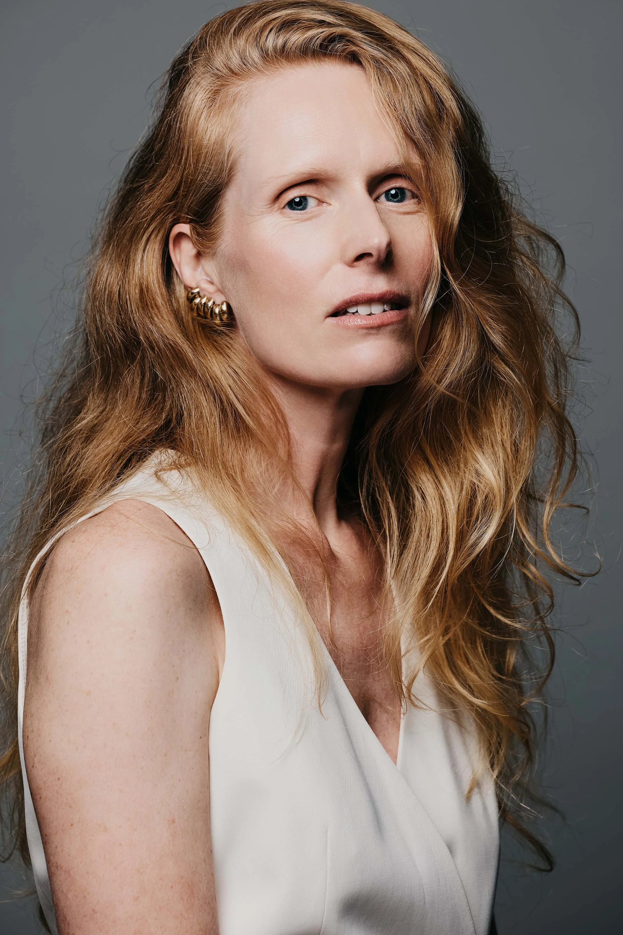 Professional headshot of Jolanda van den Berg, actress, with long auburn hair, wearing a cream sleeveless top against a gray background. Photo credit: Anne van Zantwijk.