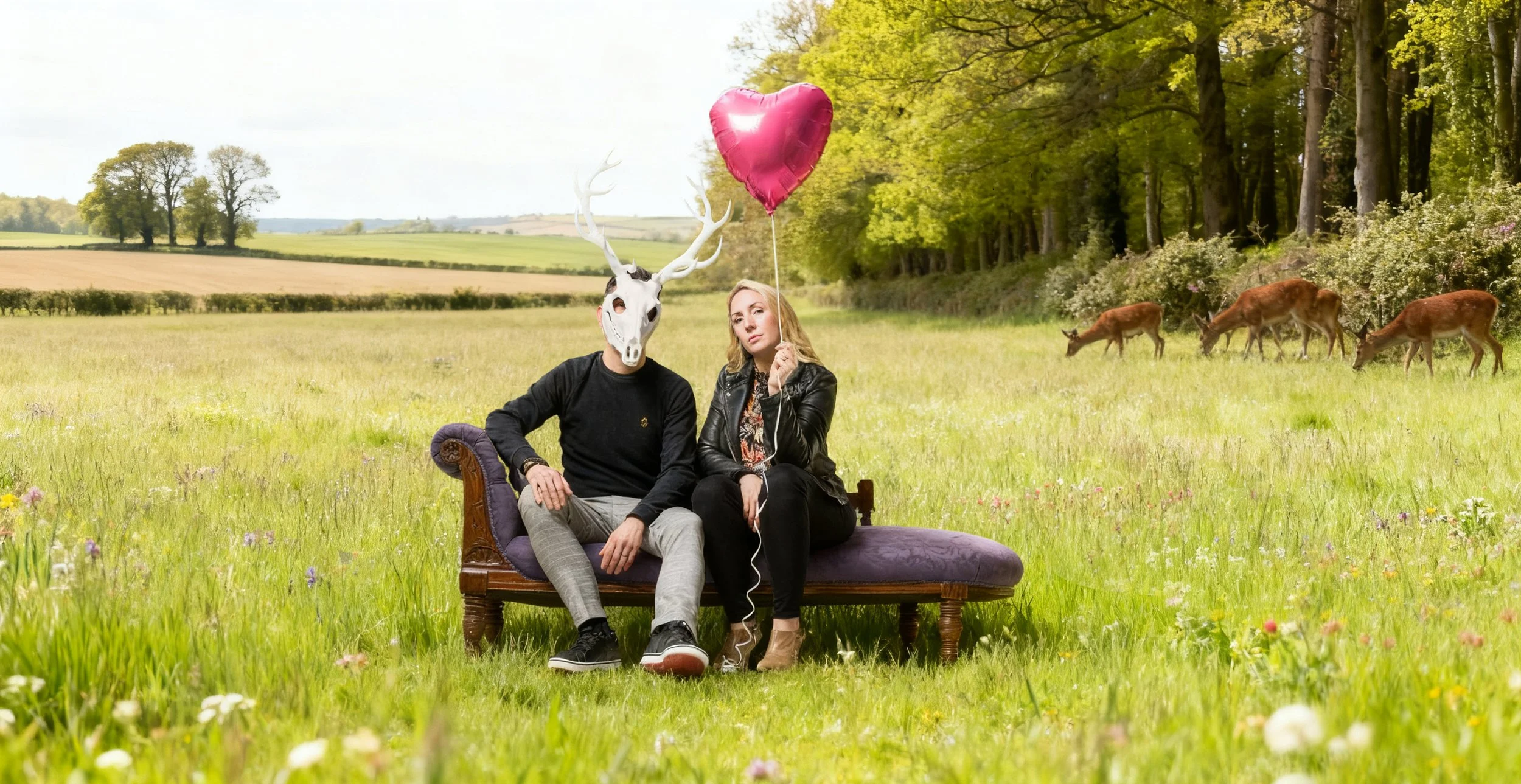 Two people sitting on a vintage chaise lounge in a meadow with trees and deer in the background; one person wears a deer mask and is holding a heart-shaped balloon, the other is holding a string attached to the balloon.