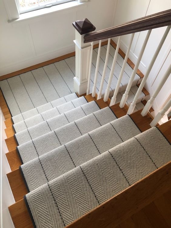 Staircase with a gray and white striped runner rug, white banister with dark brown top, wooden steps, and beige carpeted area at the top of the stairs, near a window.