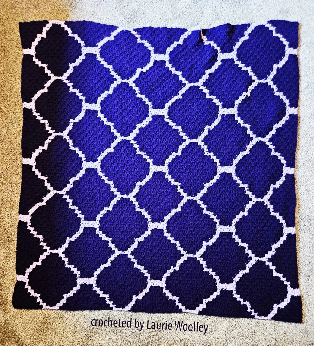 A purple and lavender quatrefoil-patterned crochet afghan draped over a neutral-colored couch. Crocheted by Laurie Woolley, showcasing the elegant and timeless quatrefoil design.