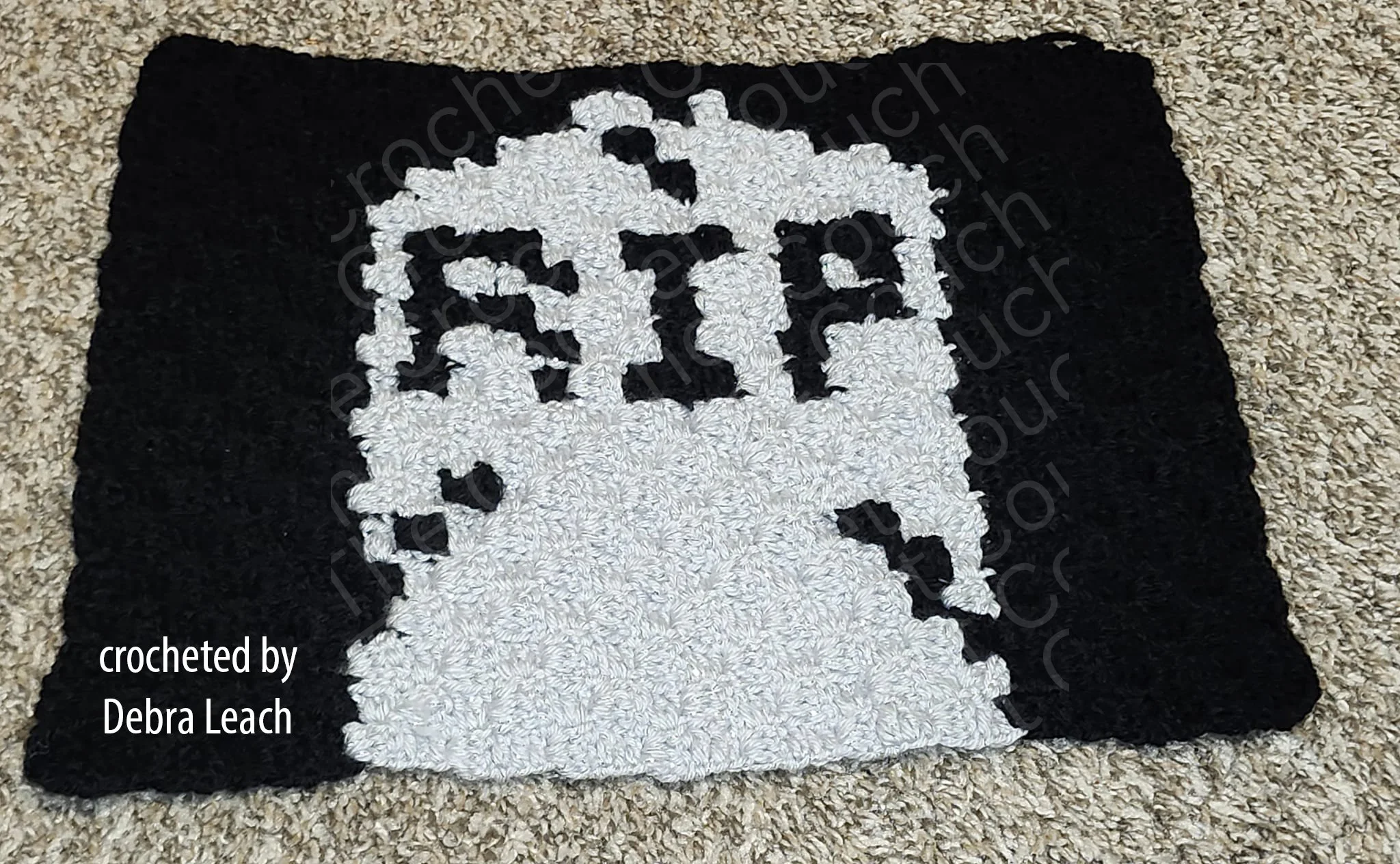 A crocheted placemat featuring a gray tombstone with the letters 'RIP' on a black background, displayed on a carpeted floor.