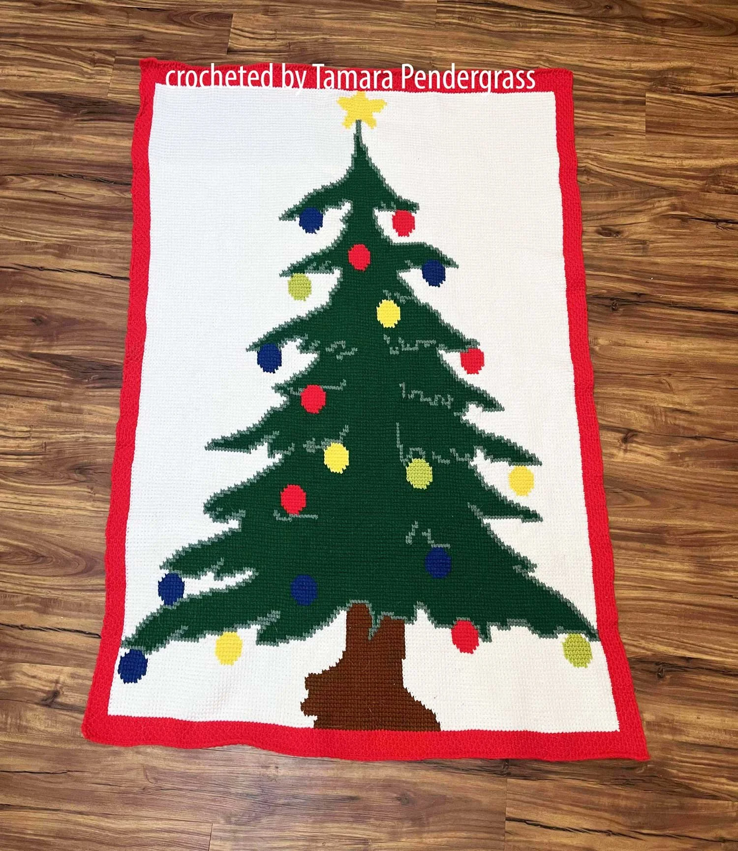 Christmas Tree Afghan featuring a large green tree with colorful round ornaments and a yellow star on top, bordered in red, displayed on a wood floor background.