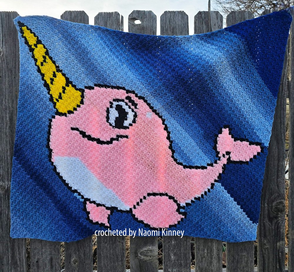 A colorful crochet Narwhal Afghan made using the corner-to-corner (C2C) technique, featuring a smiling pink narwhal with a golden horn against a blue ocean-like background.