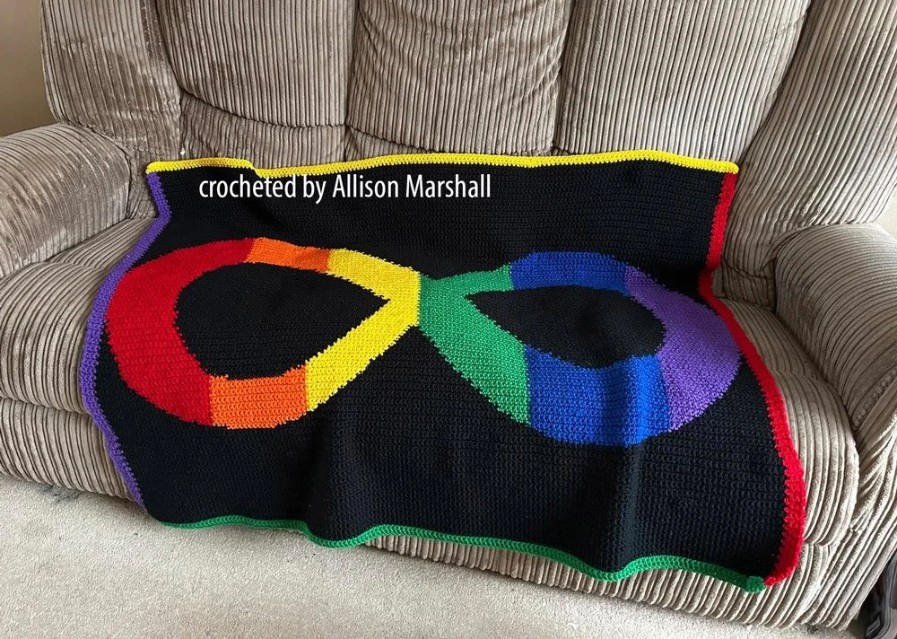 A handmade crochet afghan featuring a large rainbow infinity symbol on a black background, bordered with red, yellow, green, and purple edging.