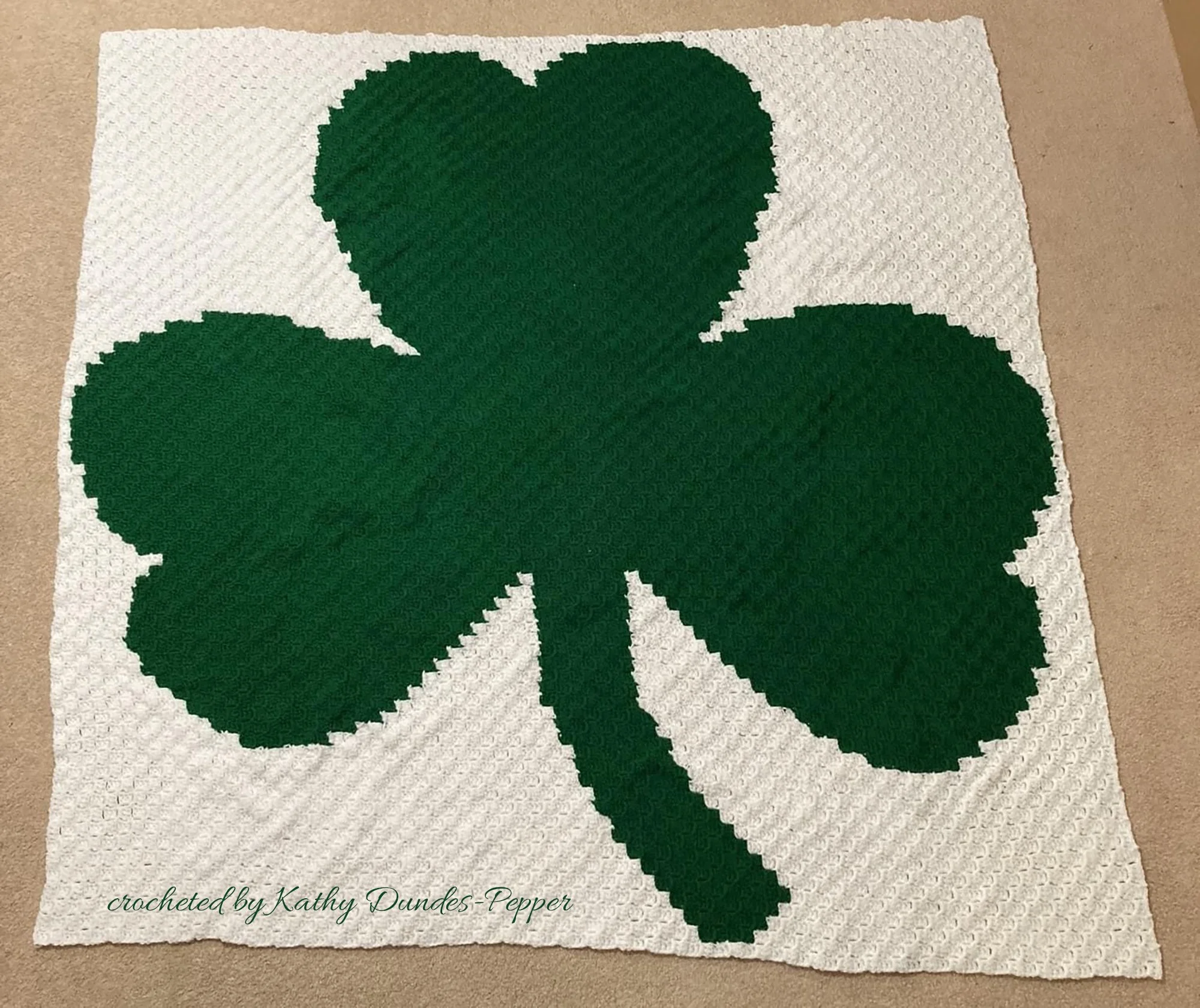 Photo shows afghan laying on the floor, it has an off white background and a green shamrock, crocheted using the C2C method by Kathy Dundes-Pepper