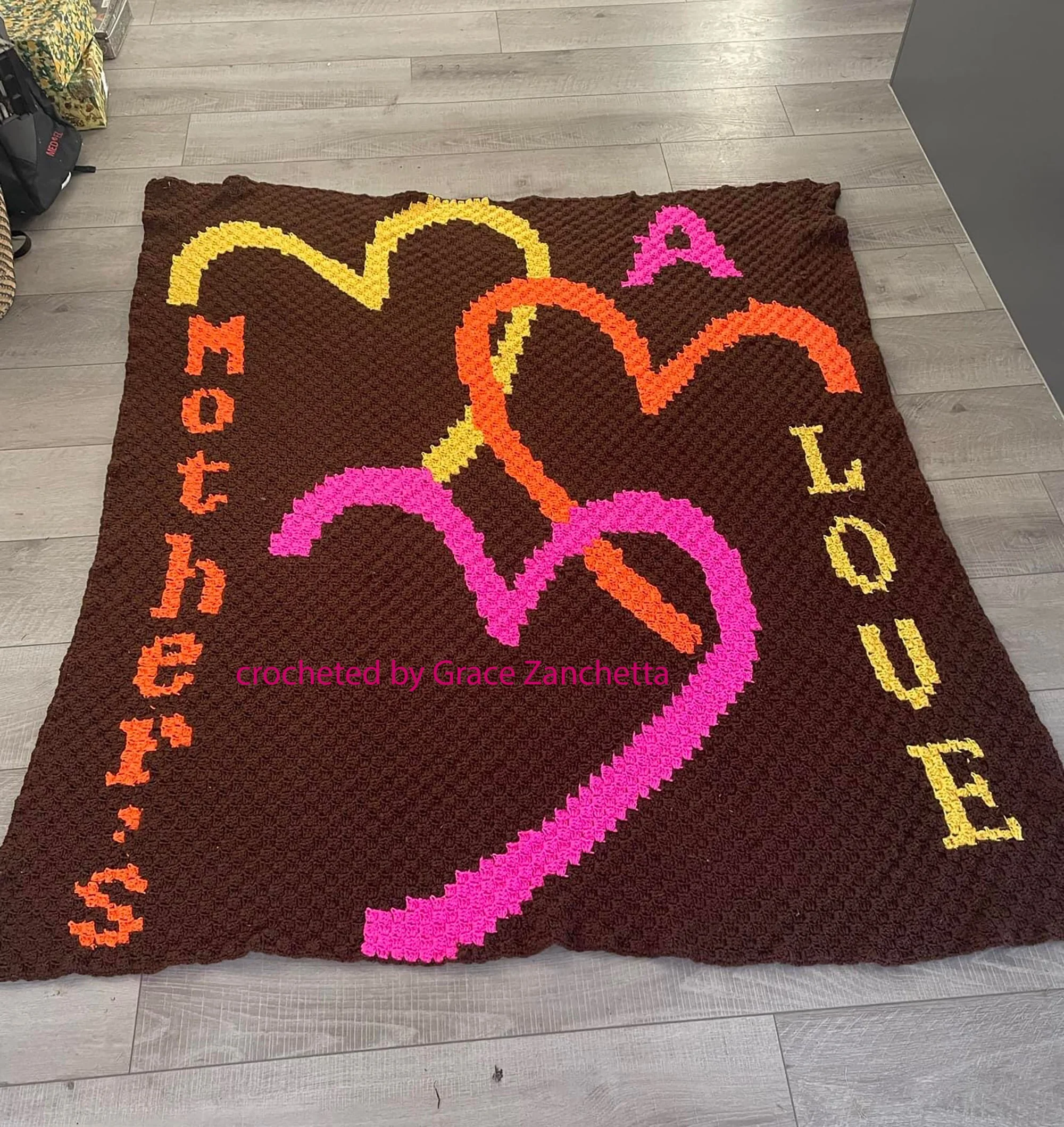 Afghan shown laid on floor, brown background, with hearts in yellow, orange and pink, all bright colors with the words A Mothers Love.  Afghan is crocheted using the C2C technique
