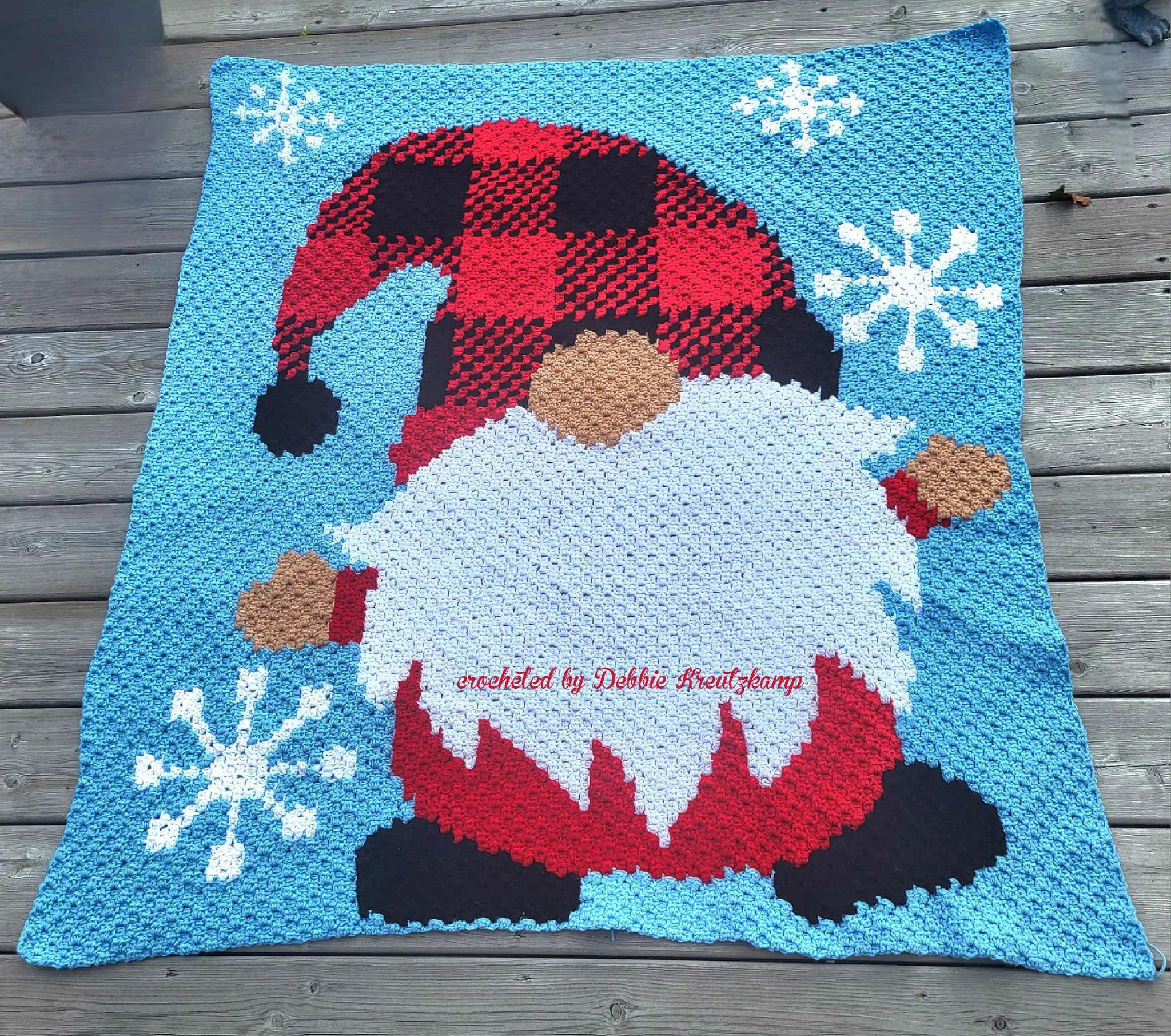 Afghan shown laid on wooden deck, blue background with snowflakes and a gnome wearing a buffalo plaid hat.  Afghan is crocheted using C2C by Debbie Kreutzkamp