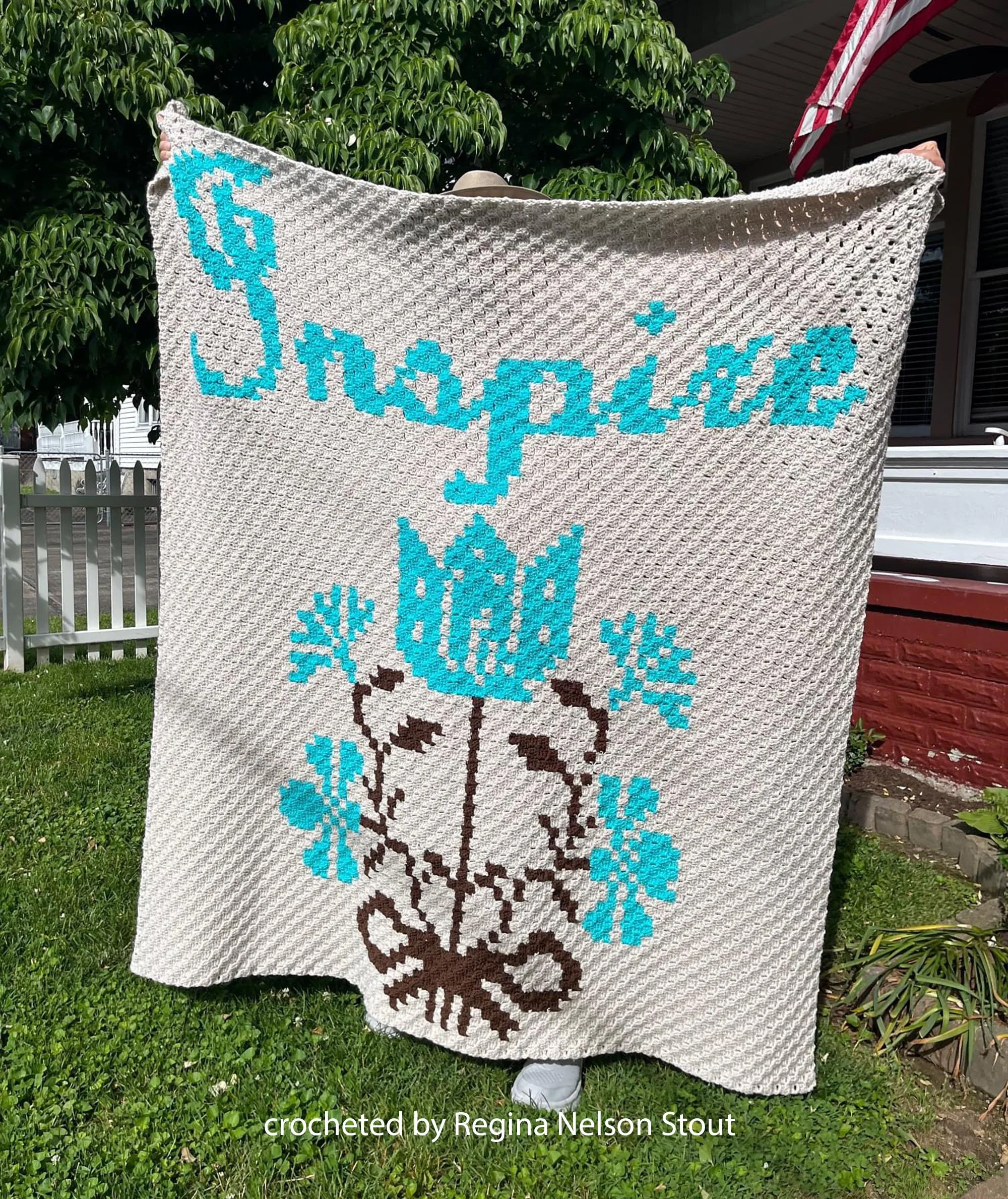 Photo shows an afghan being held up, the word Inspire is at the top in a pale teal with a teal and brown floral arrangement underneath.  The Afghan was crocheted by Regina Nelson Stout