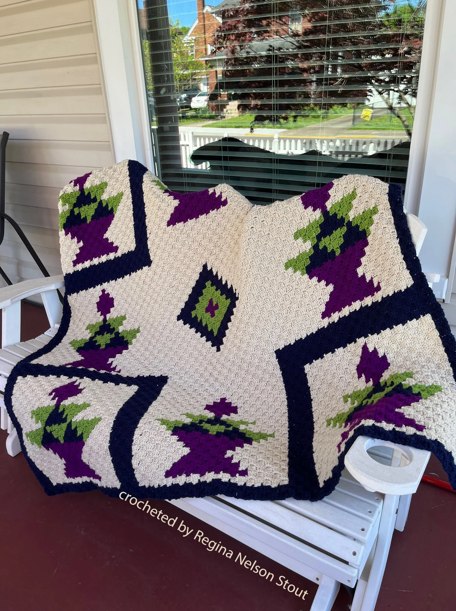 Photo shows an afghan laid on bench on front porch.  It’s a quilt pattern done in black, purple, green and off white.  The Afghan was crocheted by Regina Nelson Stout