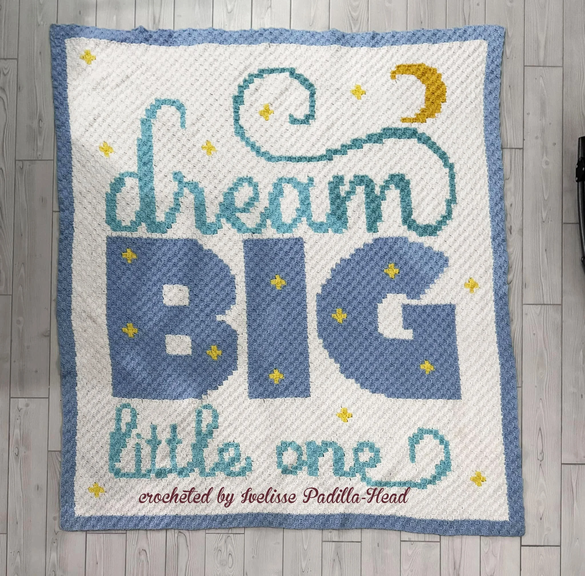 Photo shows a baby afghan laying on plank flooring - Afghan says Dream Big Little One with a moon and stars scattered throughout.  The afghan has a white background with blue, gold and teal colors