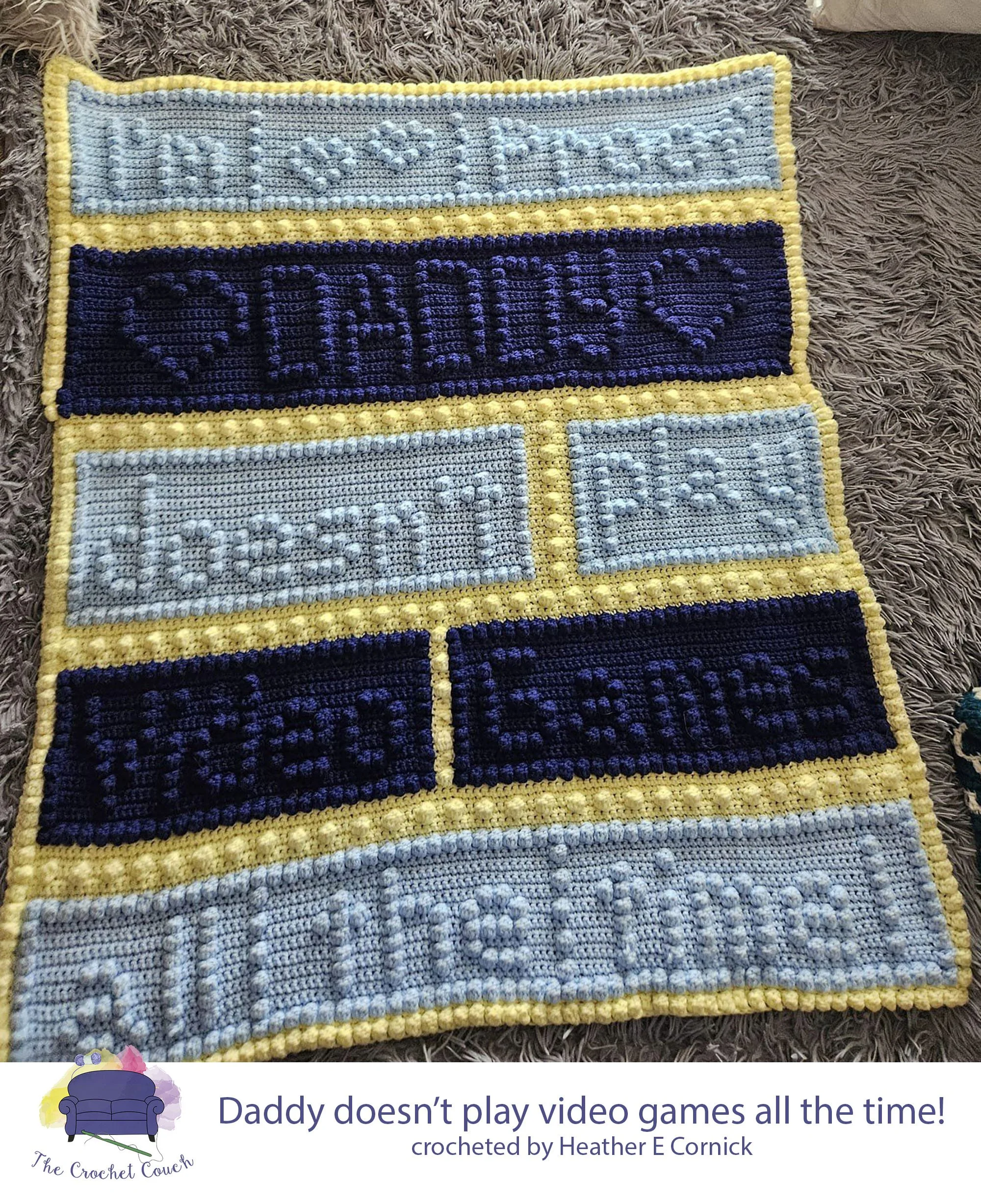 Crocheted afghan shown laying on bed - it’s done in shades of blue and yellow with single crochet and bobble stitches and says I’m Proof Daddy doesn’t play video games all the time.