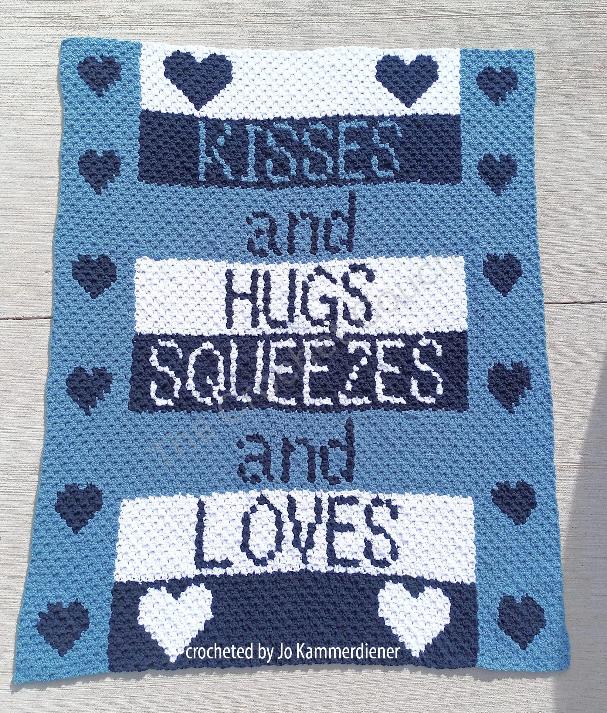 A handmade crochet afghan featuring the words "Kisses and Hugs Squeezes and Loves" in bold letters with heart designs throughout.
