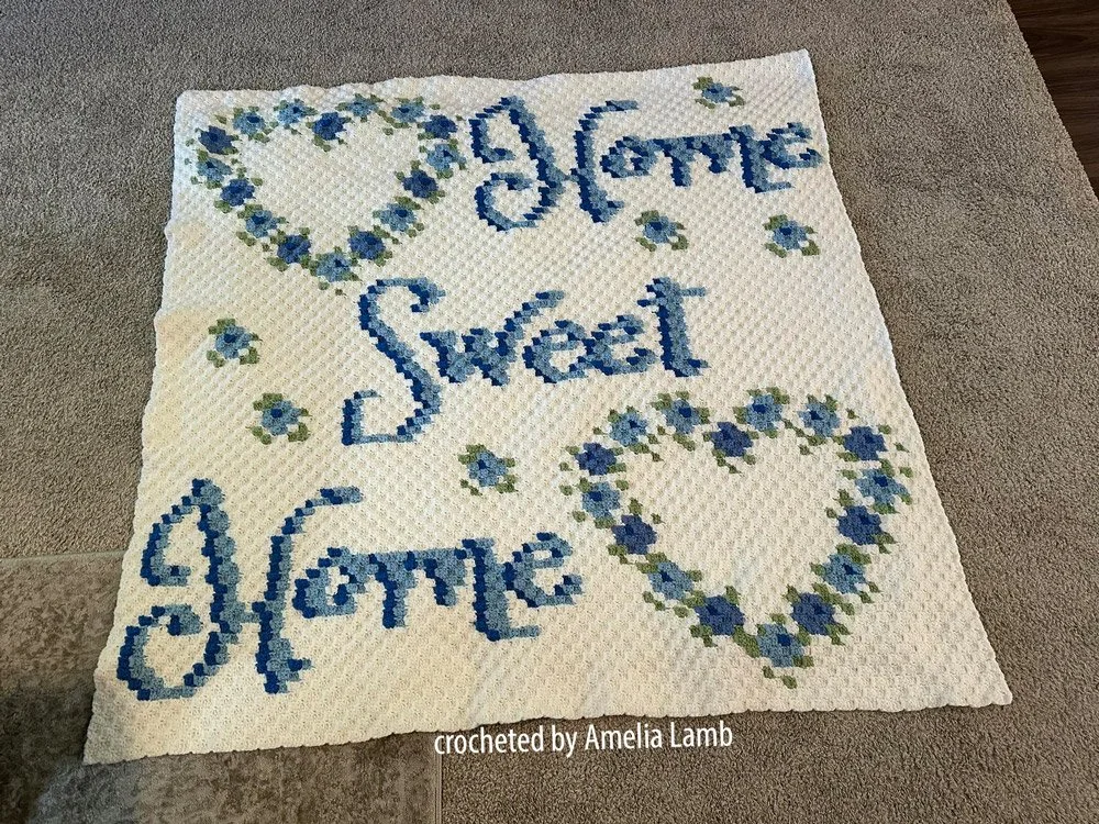 C2C crochet afghan with the phrase “Home Sweet Home” in script font, featuring two large floral hearts and scattered flowers on a cream background. Crocheted by Amelia Lamb.